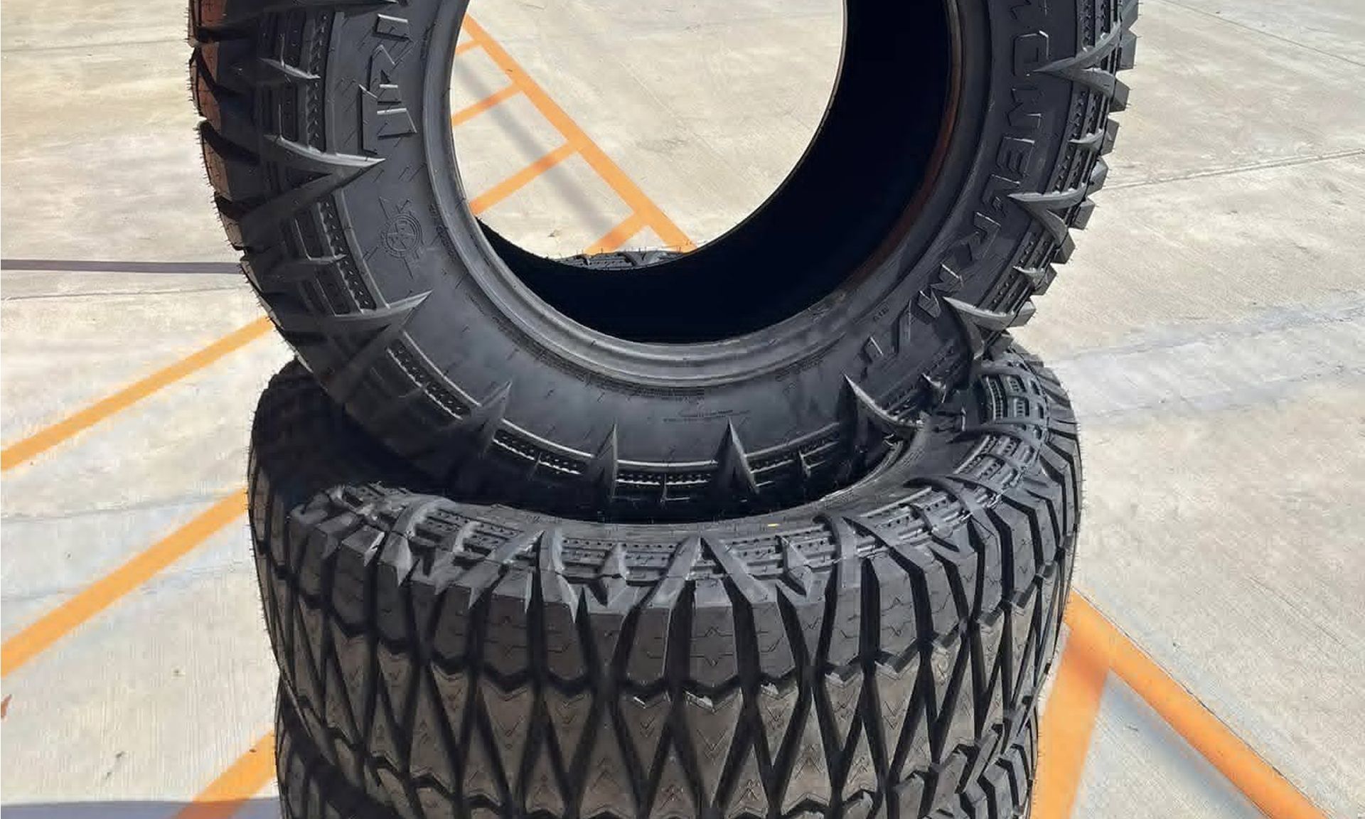 Two black off-road tires stacked on a concrete floor with orange floor markings