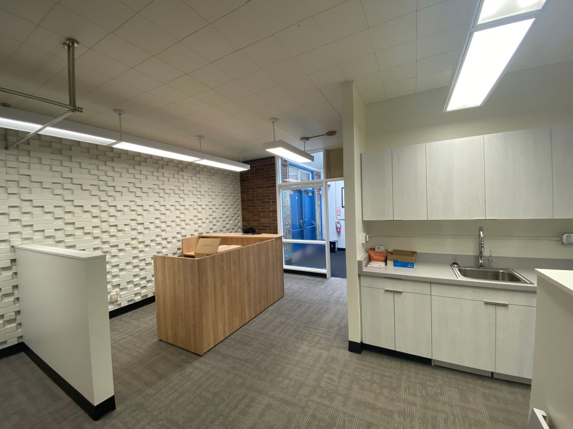 Empty modern office space with textured white wall, wooden counter, and small kitchenette.
