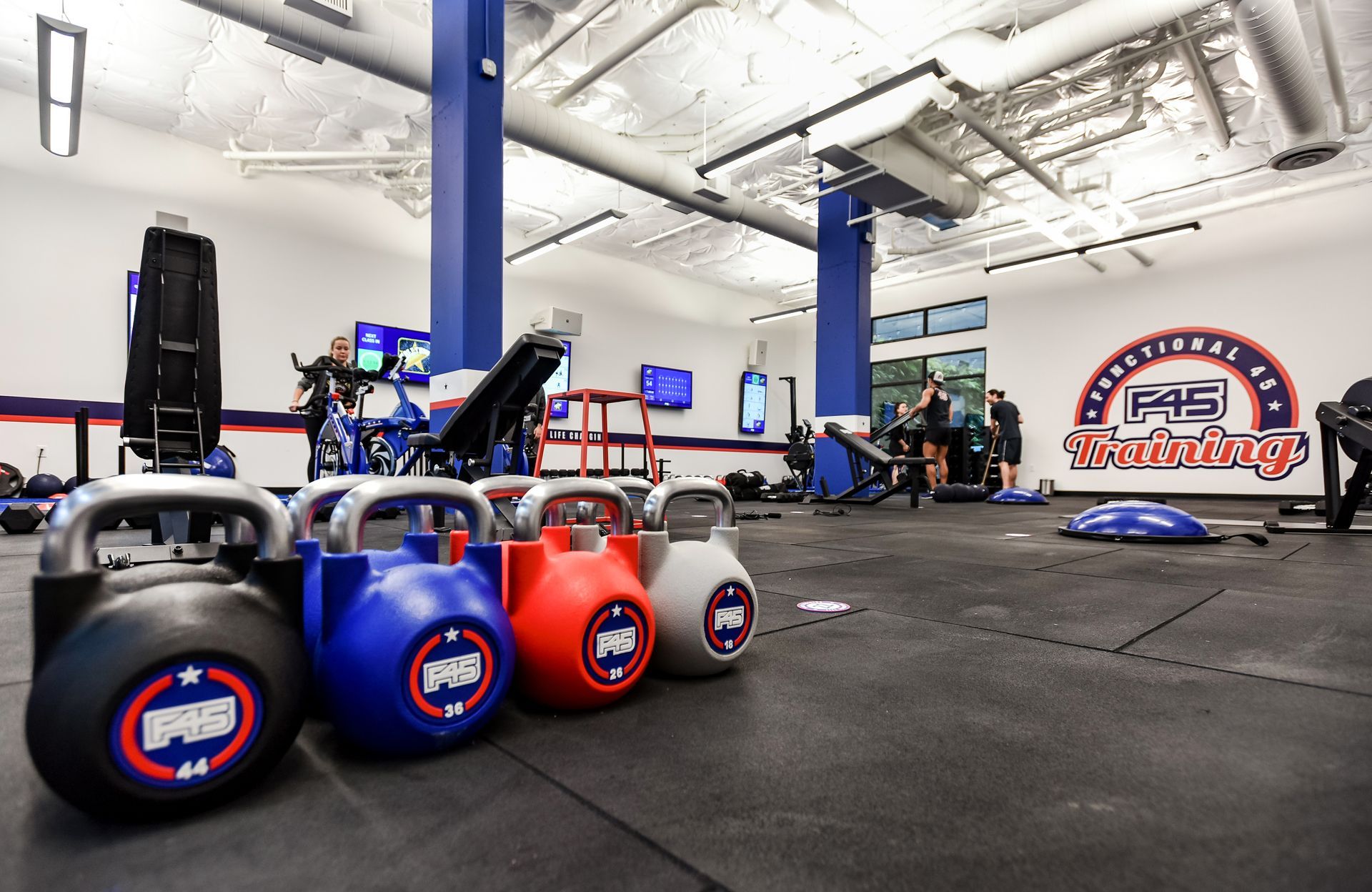 Kettlebells in a gym with people working out. Blue, red, and grey kettlebells in foreground. Exercise equipment visible.