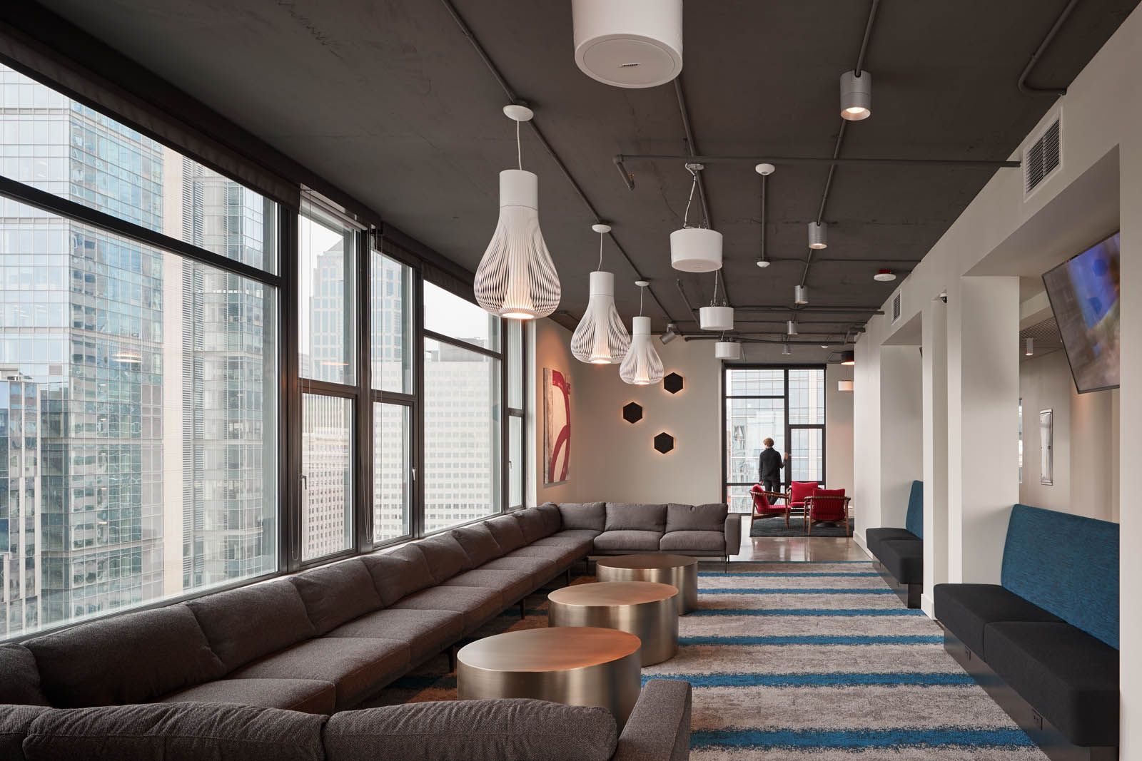 Modern lounge with large windows, long gray sofa, gold tables, and patterned rug. City views.