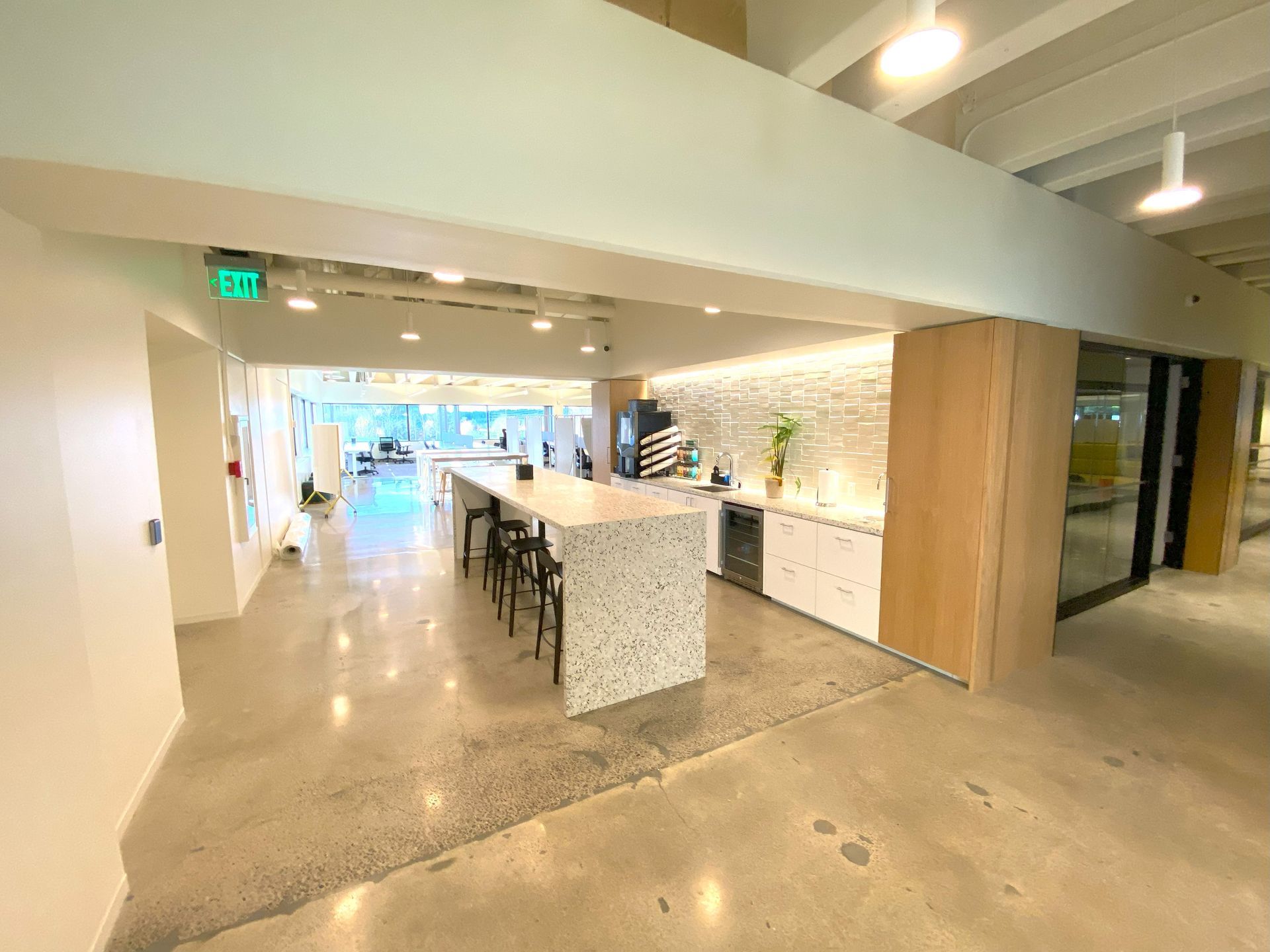 Modern office break room with concrete floors, white cabinets, island with stools, and exit sign.