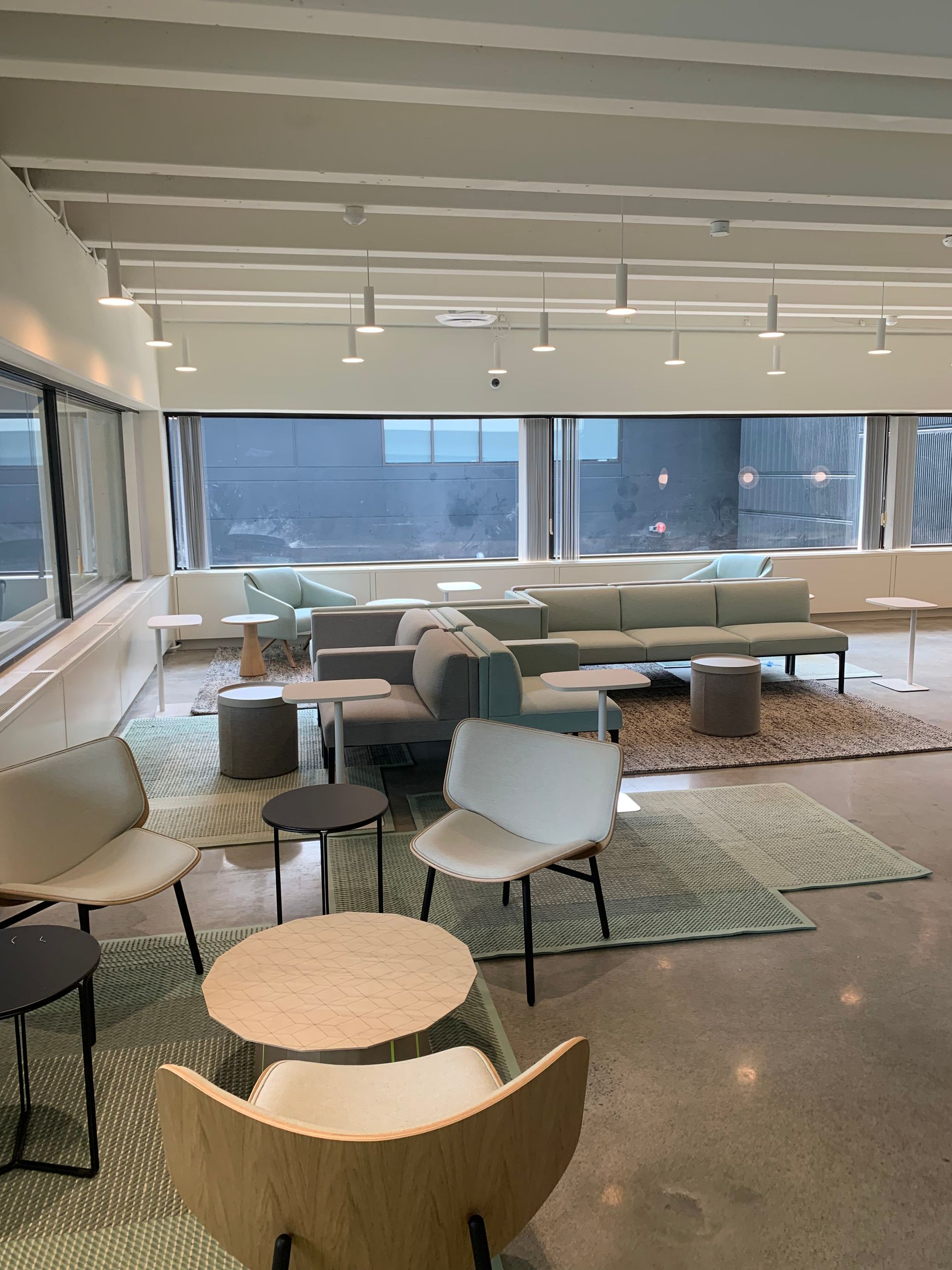 Modern lounge with couches, chairs, and tables on patterned rugs, lit by overhead lights and a large window.