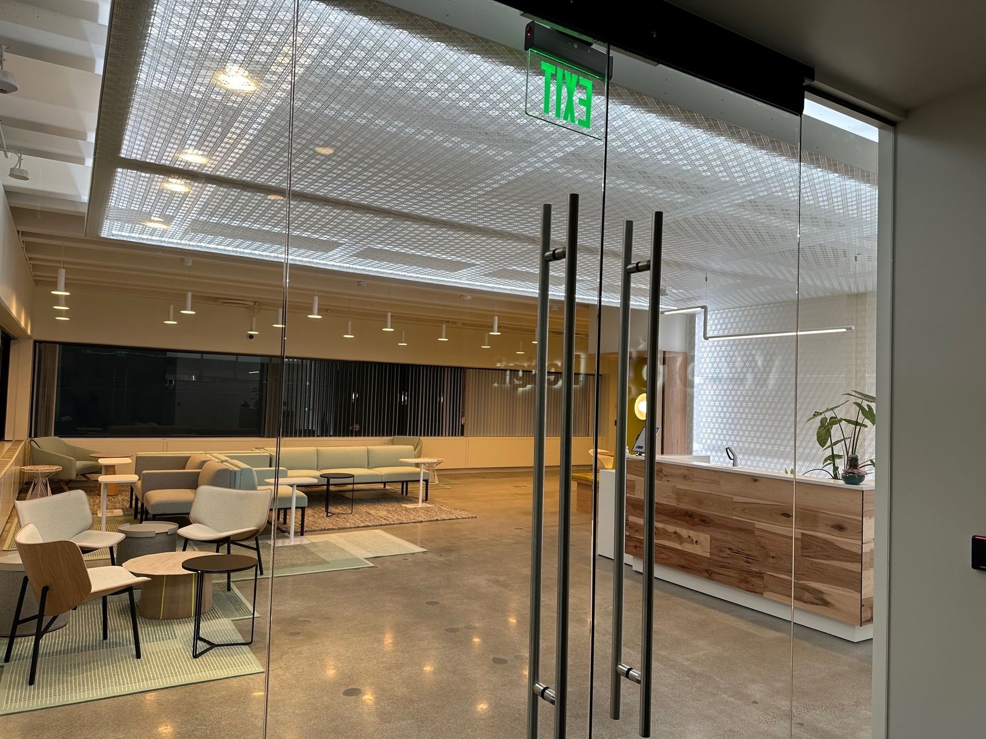 Glass entry door to modern office reception area. Gray concrete floor, textured ceiling, light wood reception desk, seating area.