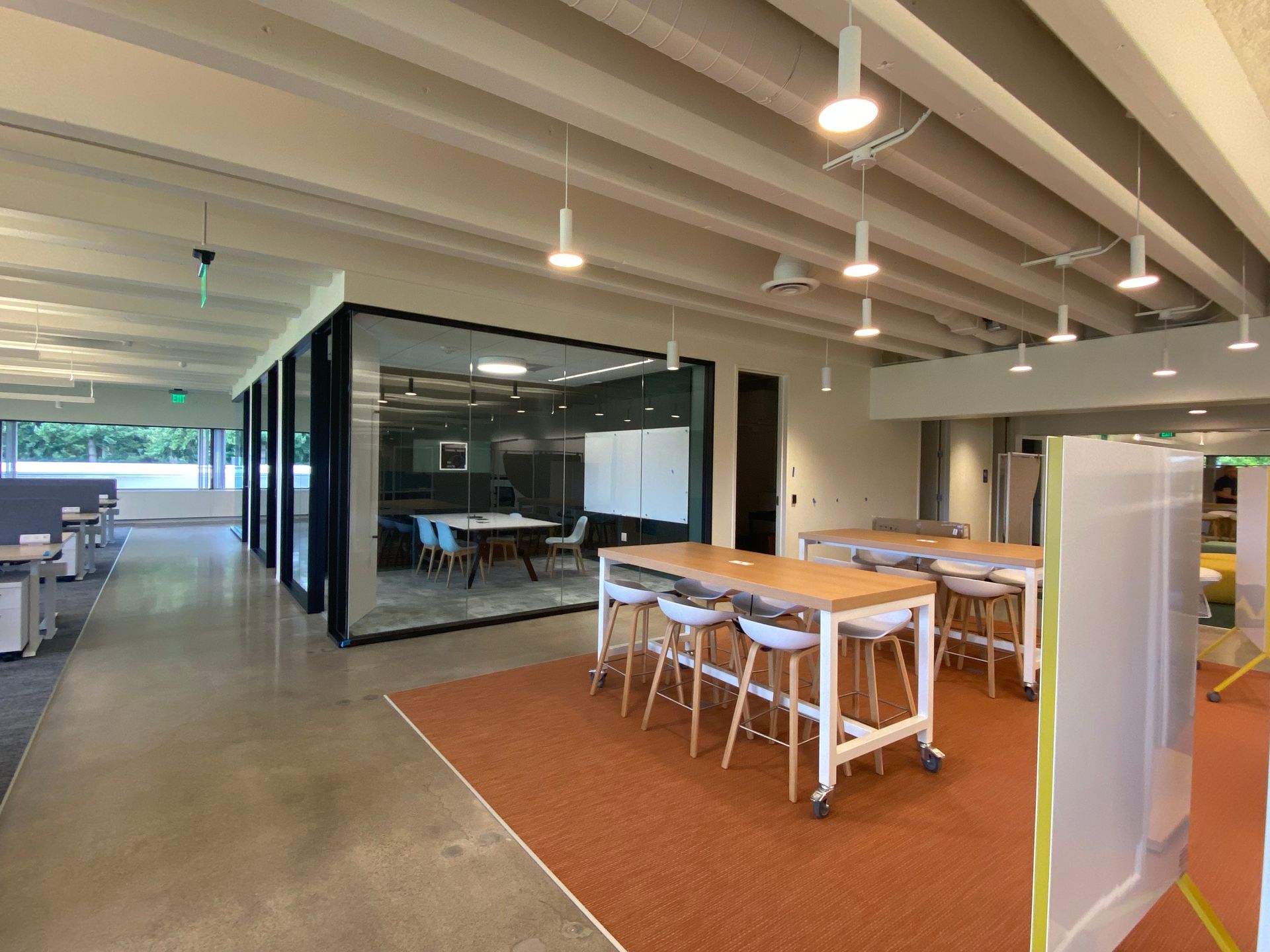 Modern office space with a glass-walled conference room, communal tables, and exposed ceiling beams.
