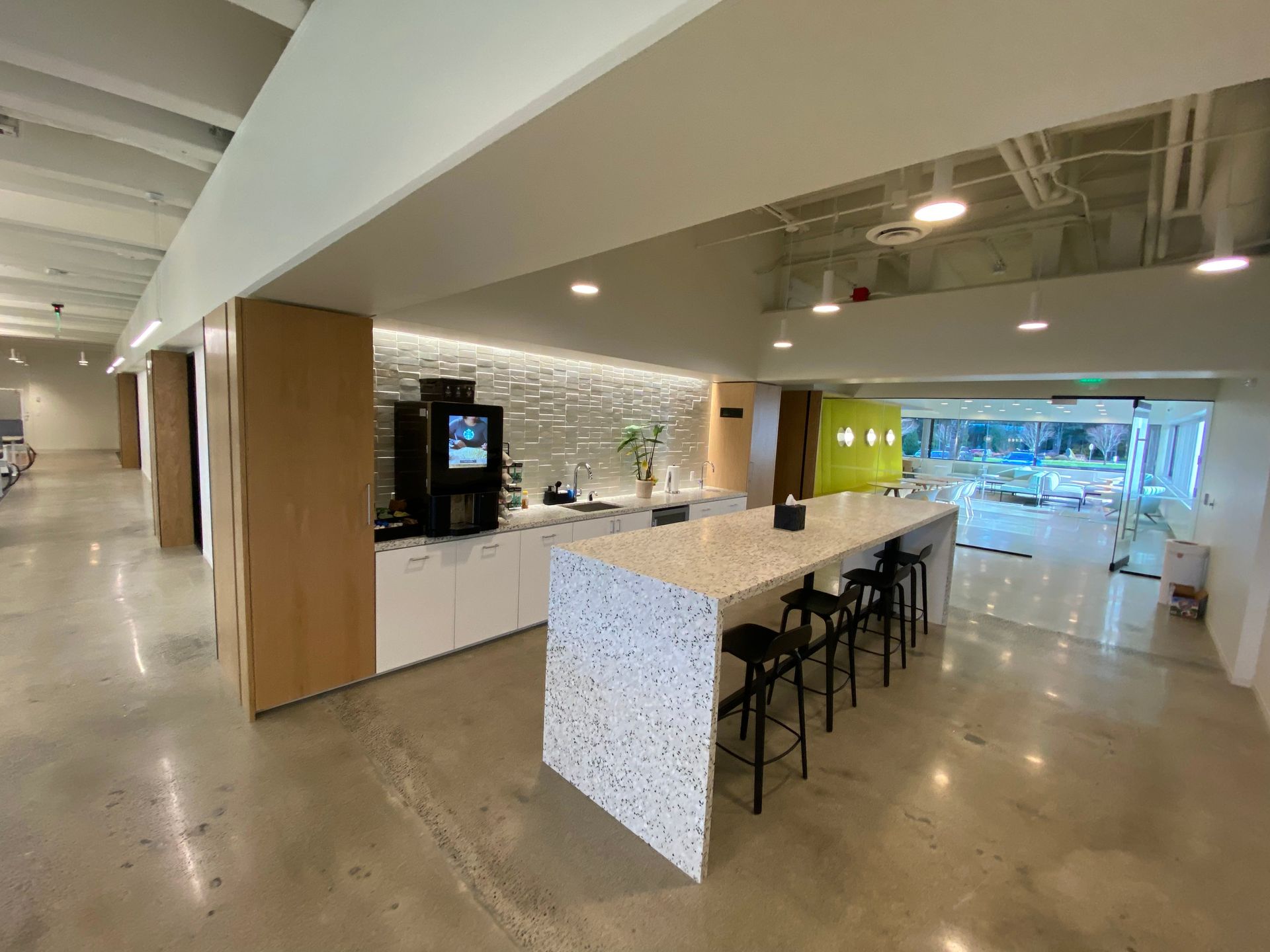 An office break area with a coffee machine, a long counter with stools, and a bright green accent wall.