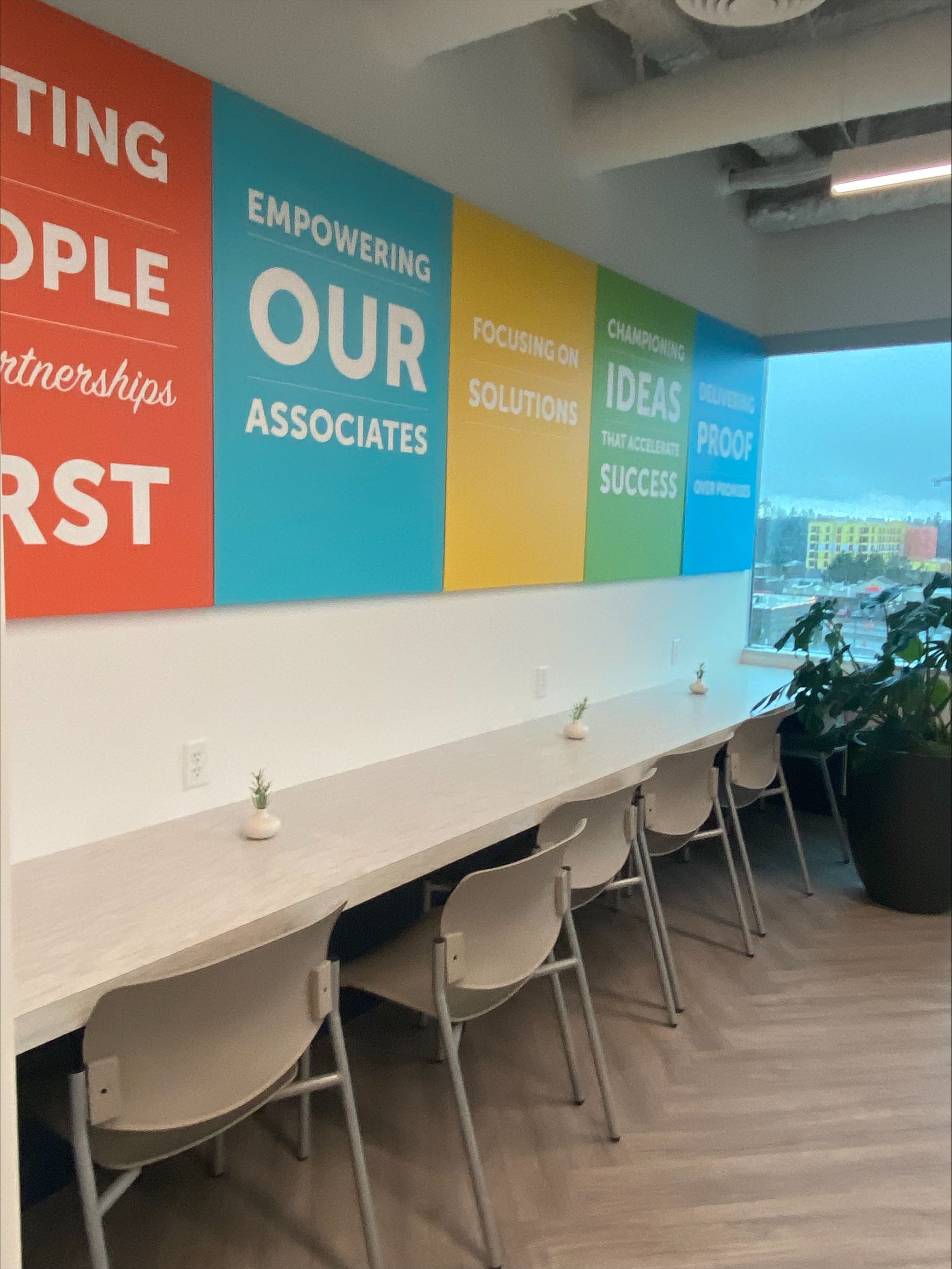 Long desk with chairs against a wall with colorful motivational words. Plants and a window are to the right.