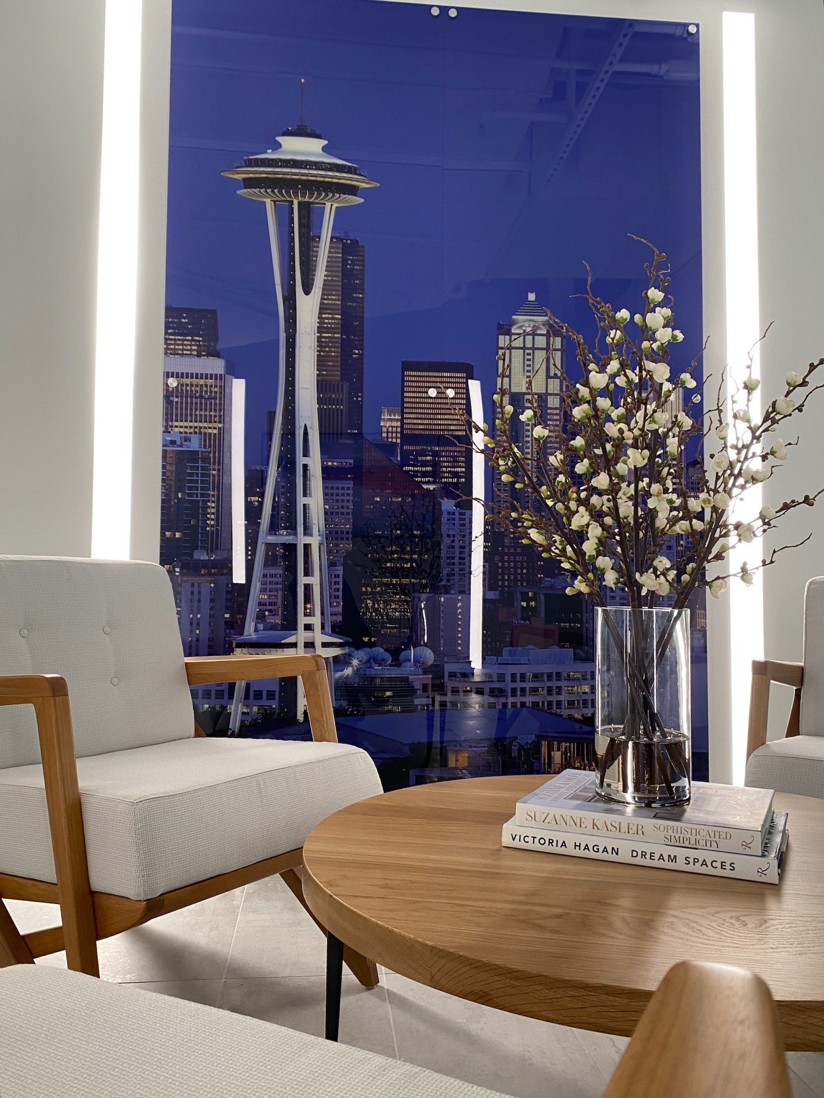 Living room with Seattle skyline view; white chairs, wooden table with flowers and books.