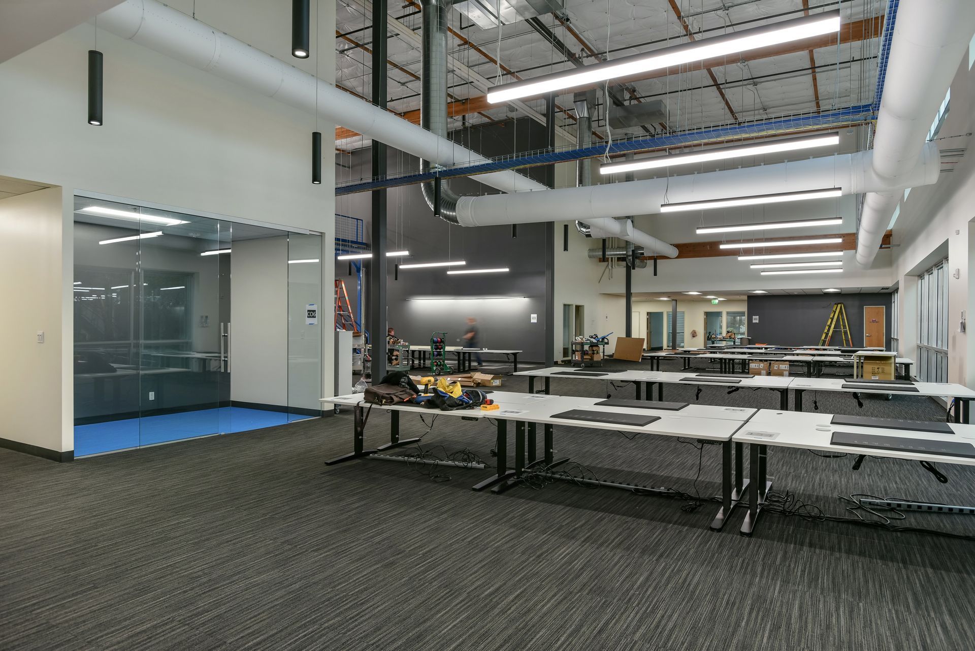 Open-plan workspace with multiple tables, blue carpeted area, glass-walled room, and exposed ceiling with lighting and ductwork.