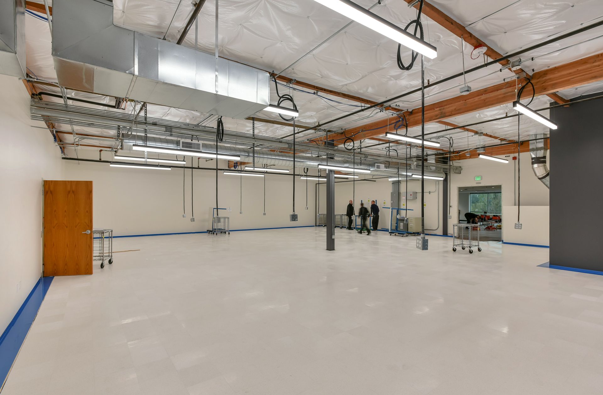 Brightly lit, empty warehouse space with exposed beams and ductwork. Three figures stand near the center.