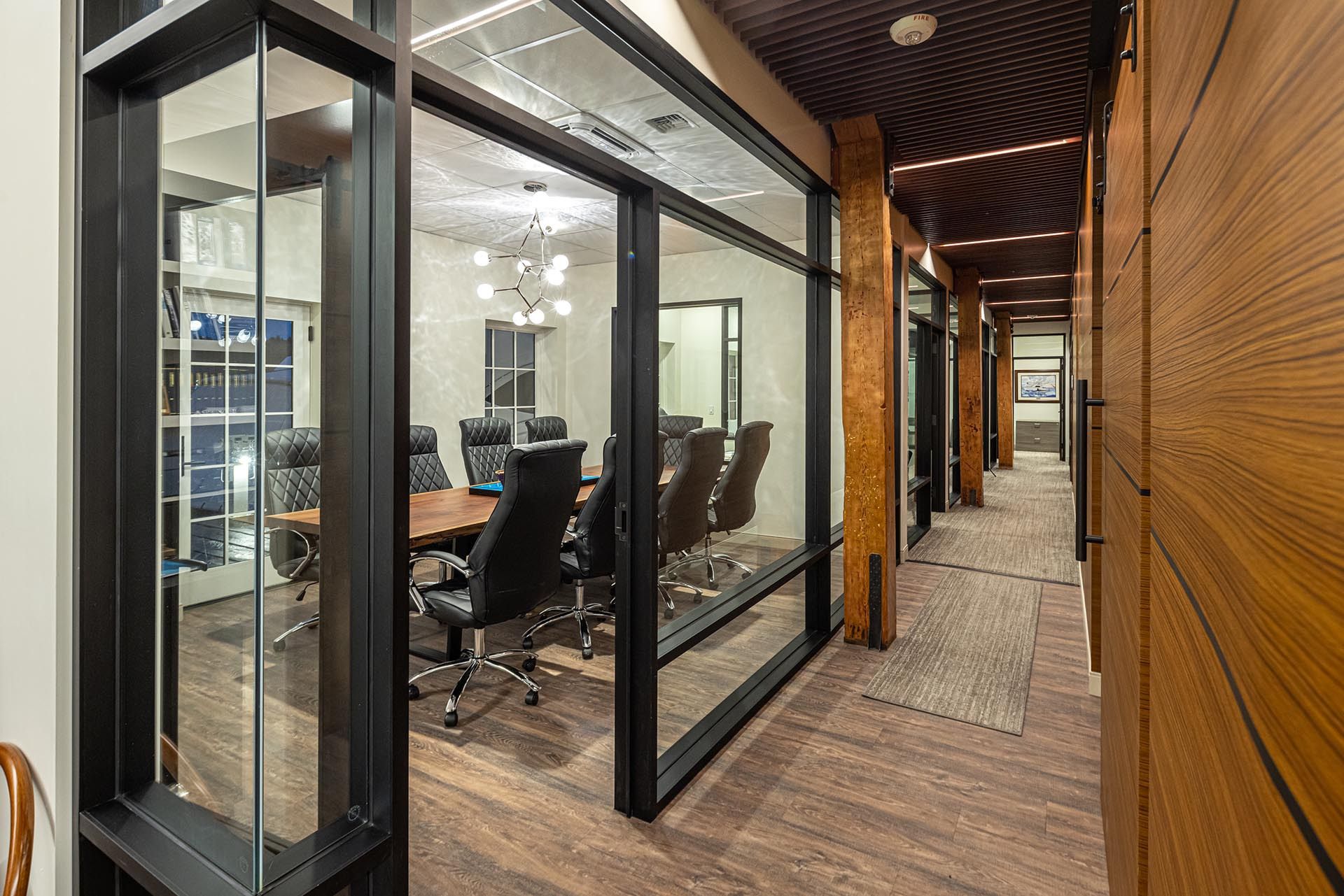 Hallway with glass-walled conference room visible. Black chairs surround a wooden table. Warm wood paneling and a patterned ceiling.