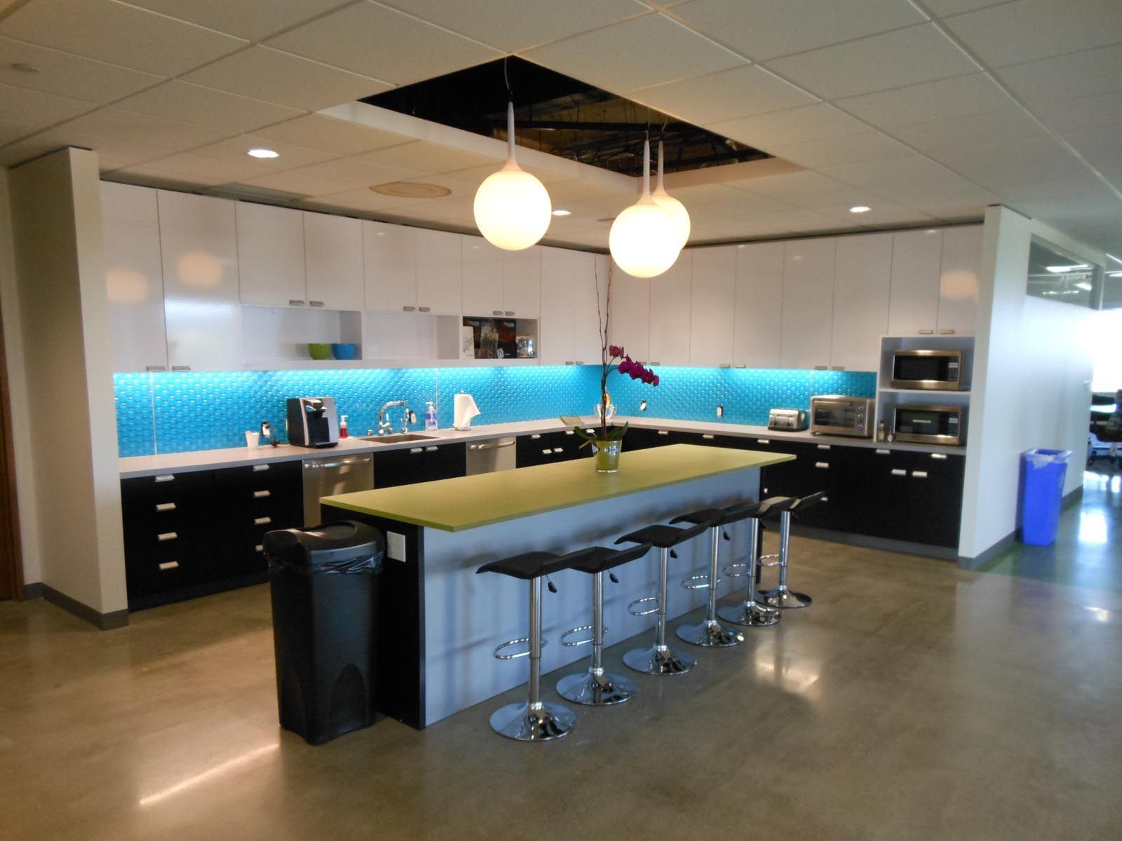 Modern office kitchen with white and black cabinets, turquoise backsplash, island with stools, and globe pendant lights.