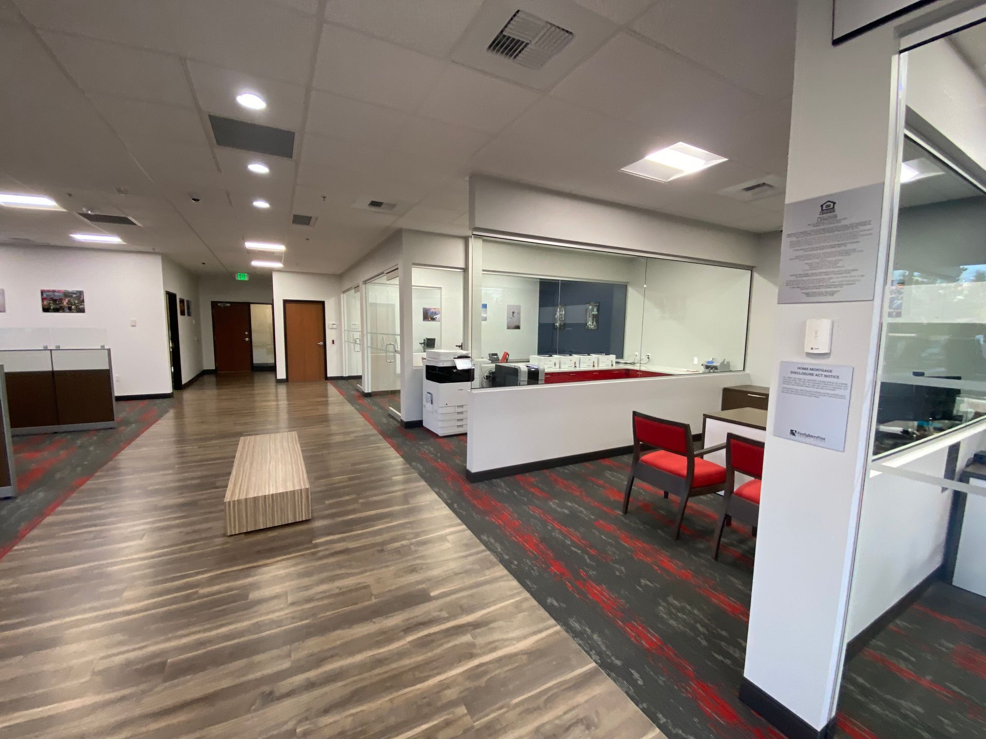 An office hallway with wood-look flooring, a bench, and glass-walled offices.