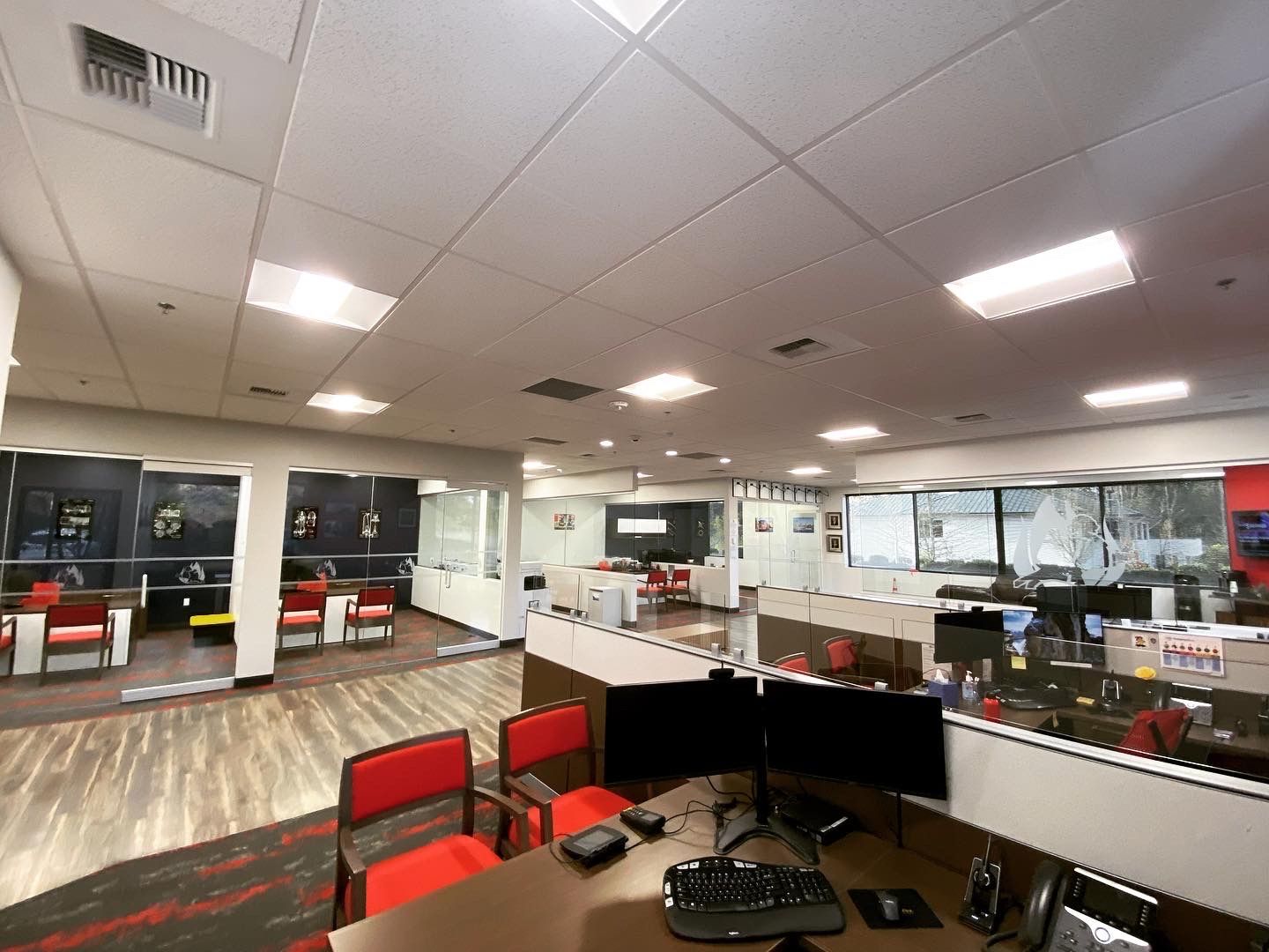 Interior view of an office with cubicles, desks, and red chairs. Fluorescent lighting illuminates the space.