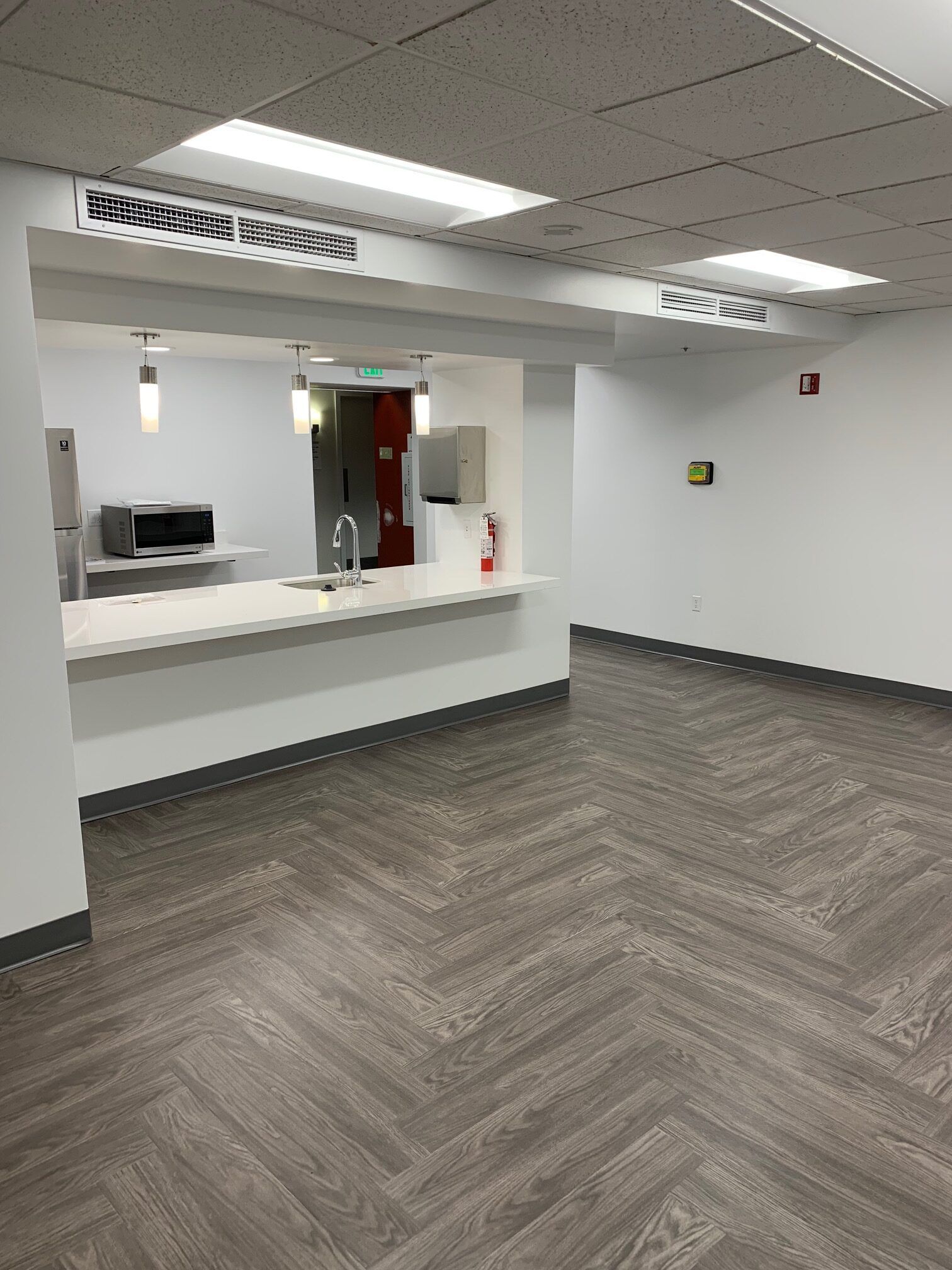 Empty room with herringbone pattern flooring and a kitchen area visible.