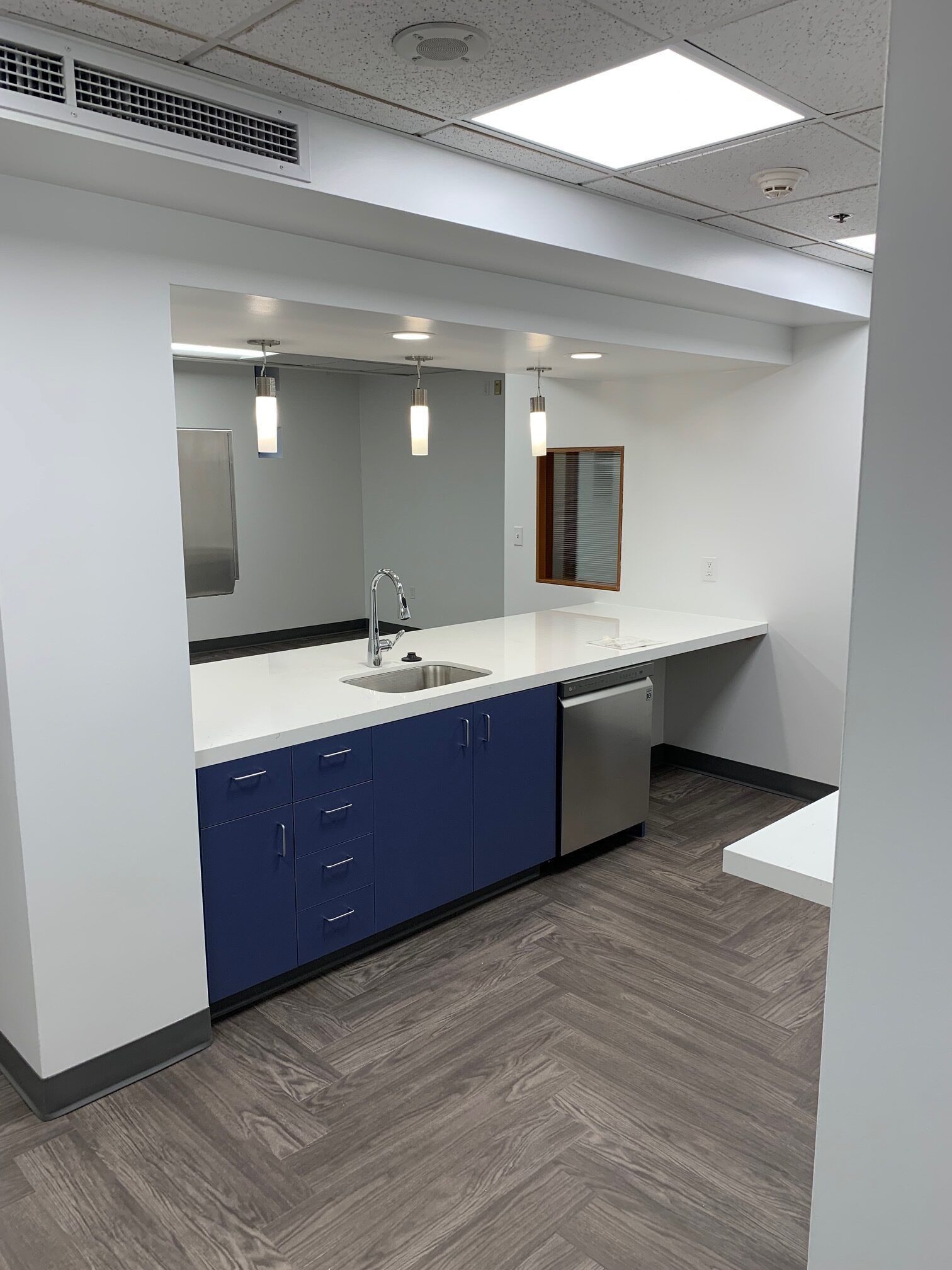 Modern office kitchenette with blue cabinets, white countertop, stainless steel sink and appliances, and wood-look flooring.