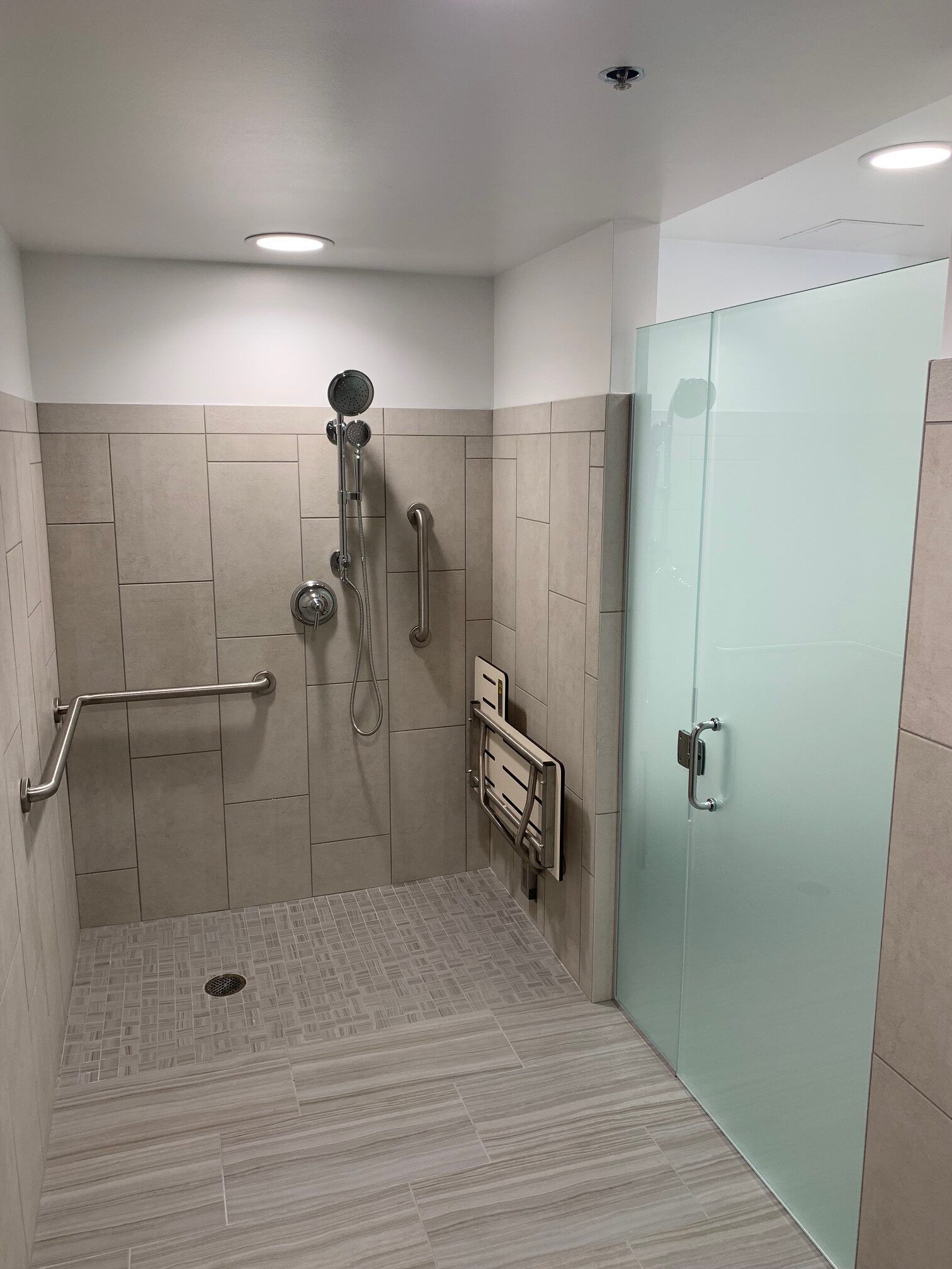 Accessible shower with grab bars and fold-down seat, beige tile walls, frosted glass door.