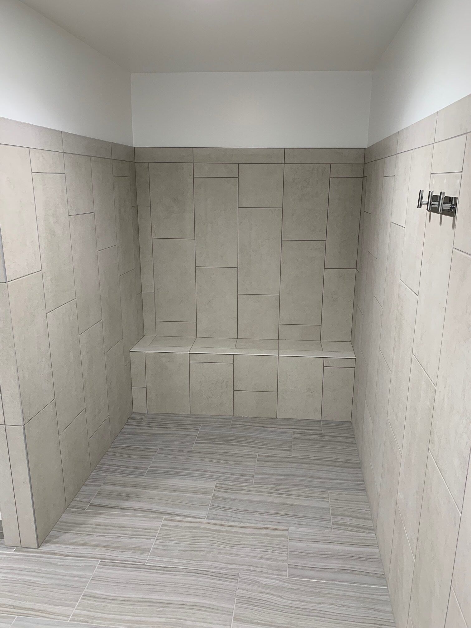 Shower stall with light-colored textured tiles, a built-in bench, and shower fixtures on the wall.