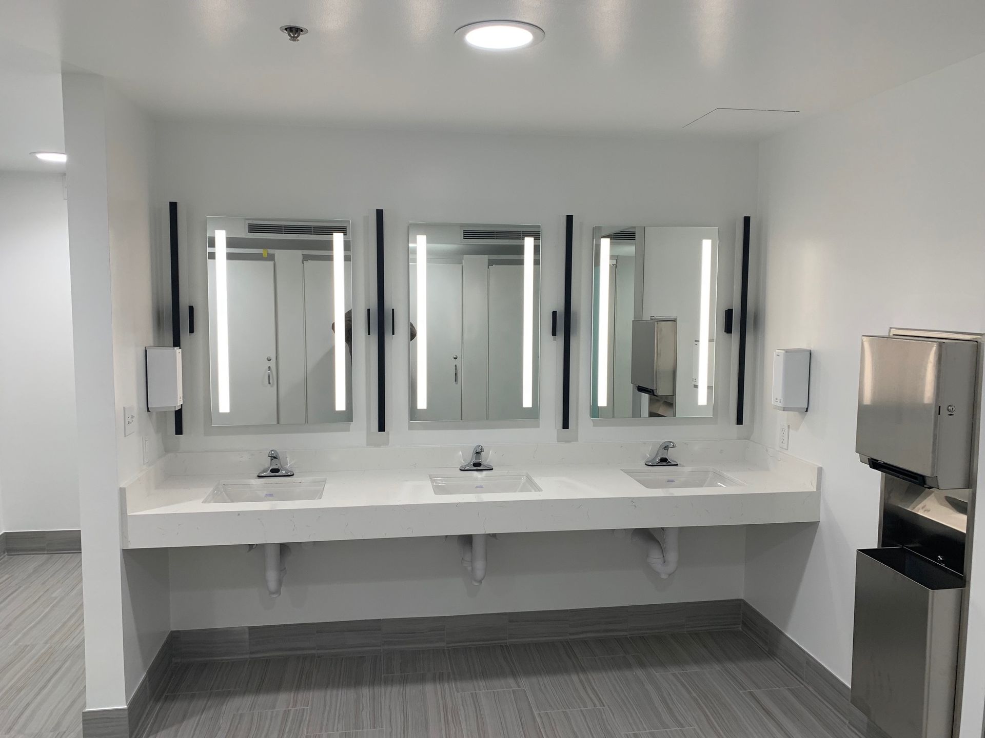 Public restroom with three sinks, mirrors with lights, and paper towel dispenser.