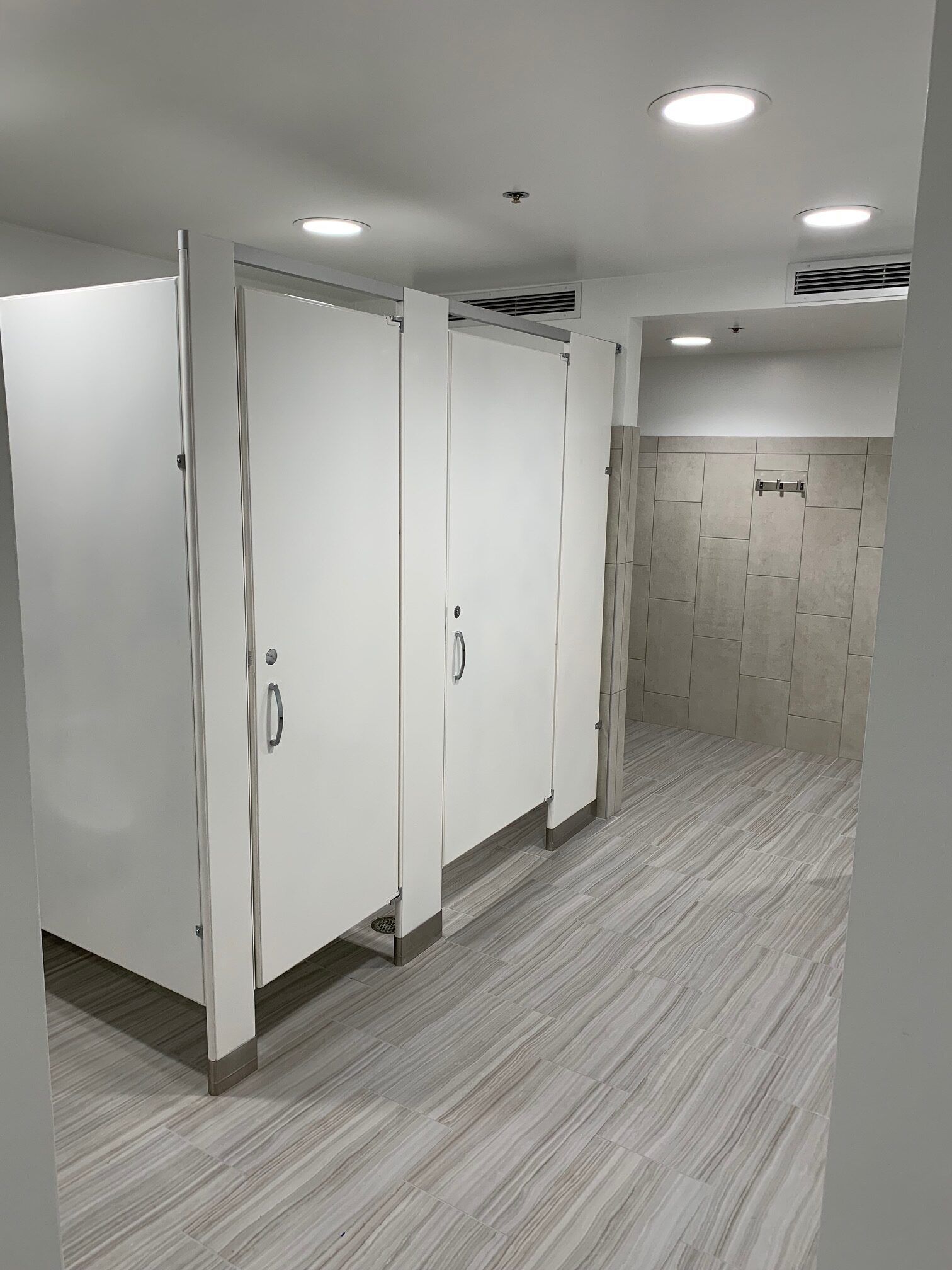 Bathroom stalls, white doors, gray tiled floor and walls, overhead lighting.