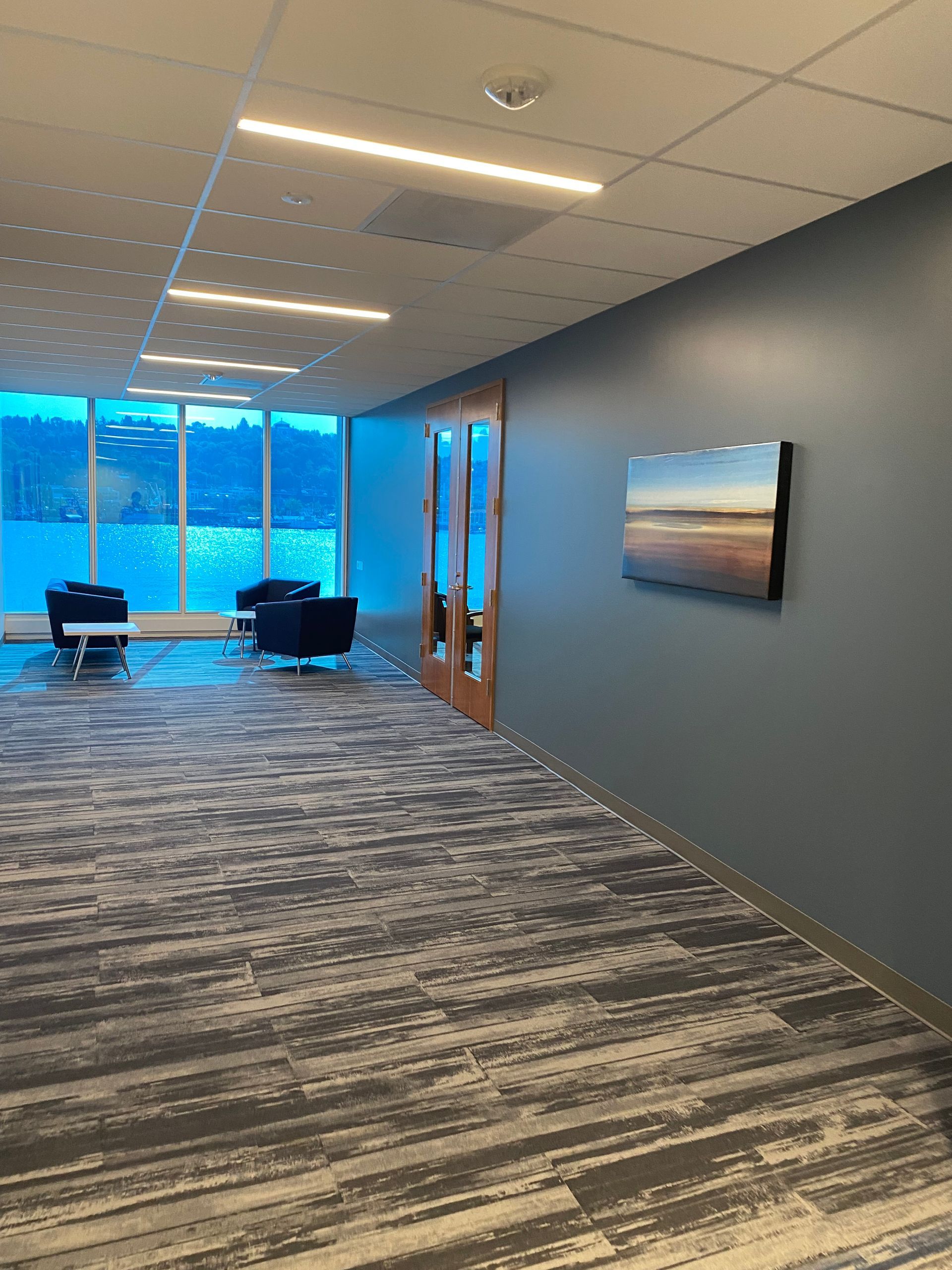 Office hallway with blue walls, carpet, and windows. Artwork on the wall. Several chairs in a seating area.