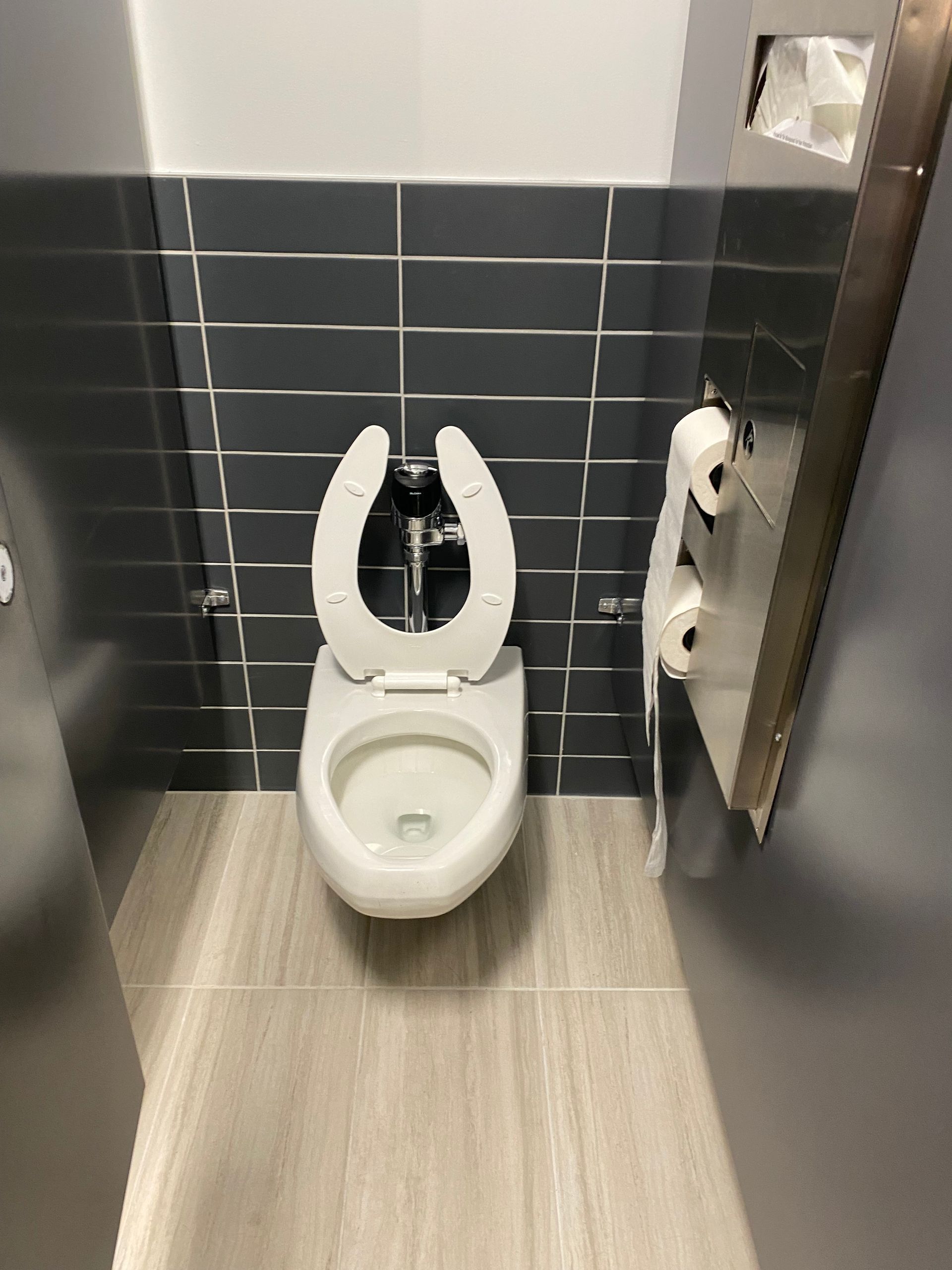Public restroom stall with toilet, gray tile wall, and stainless steel door.