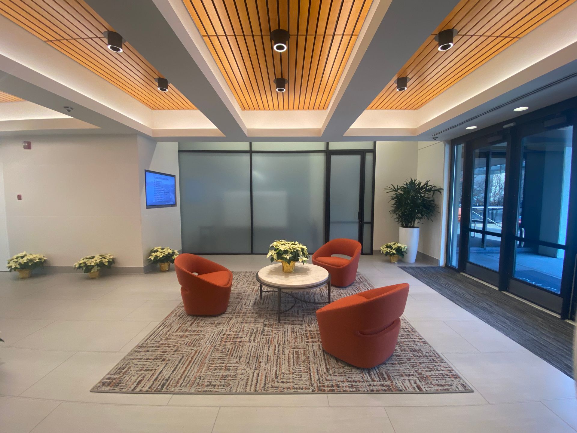 Lobby with orange chairs, a coffee table, and plants. Wooden ceiling and glass doors.
