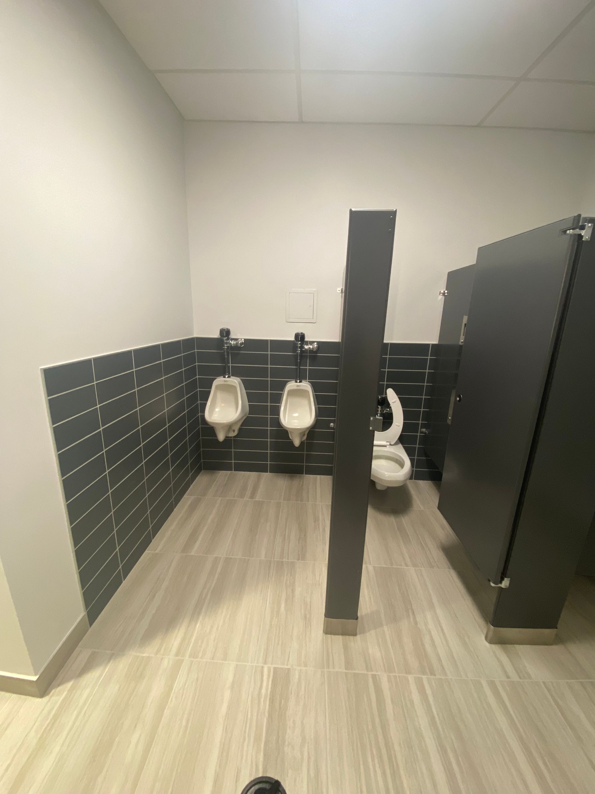 Public restroom with two urinals and a toilet stall, gray tile and light wood-look floor.