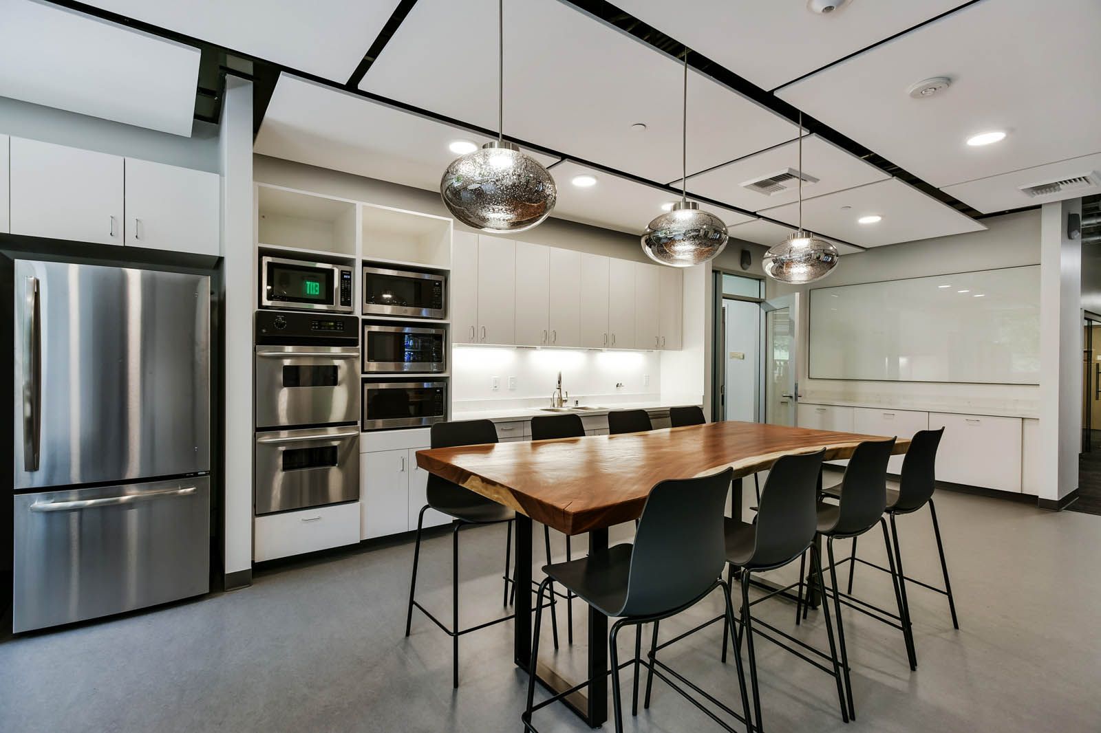 Modern office kitchen with stainless steel appliances, long wooden table, and black chairs.