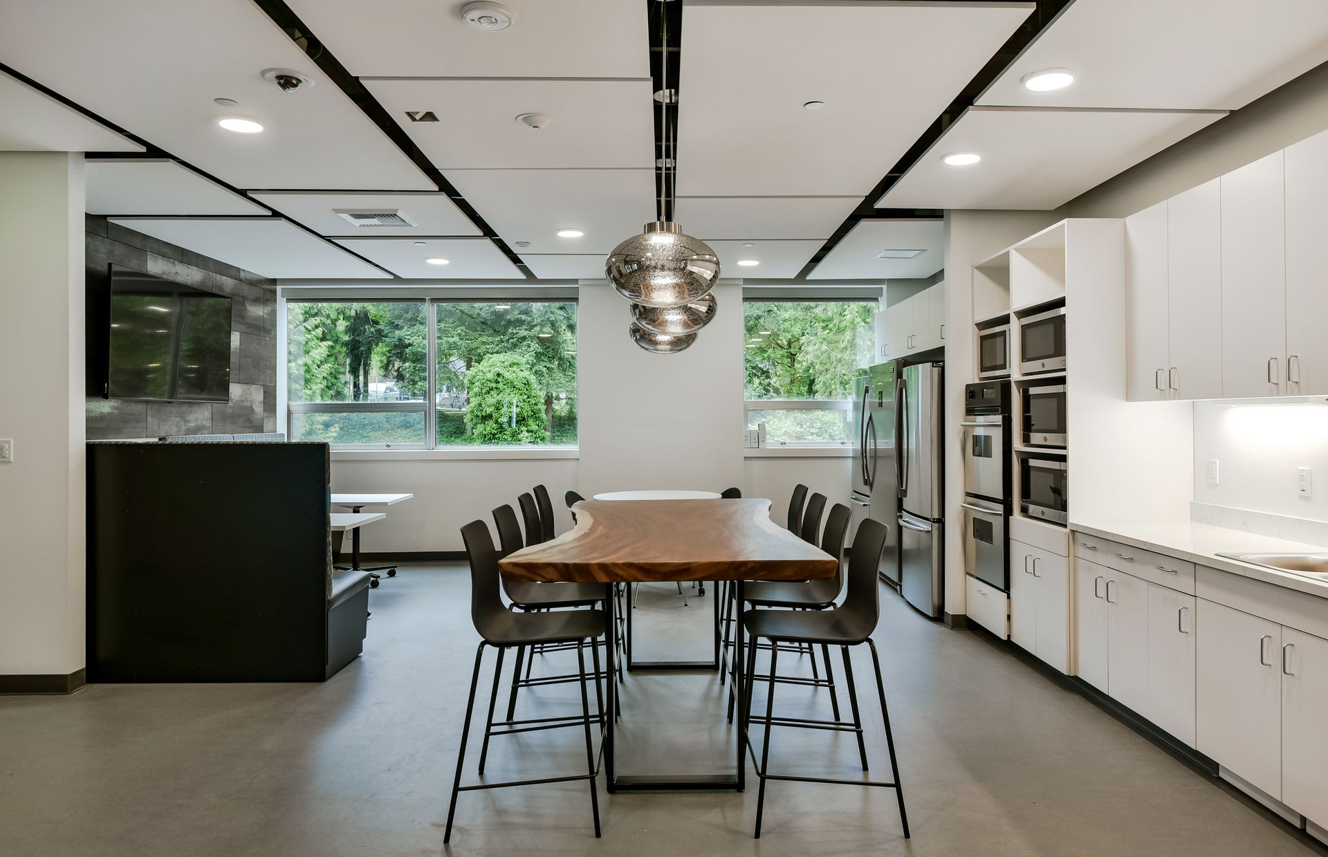 Modern office kitchen with long wooden table, black chairs, white cabinets, and stainless steel appliances.