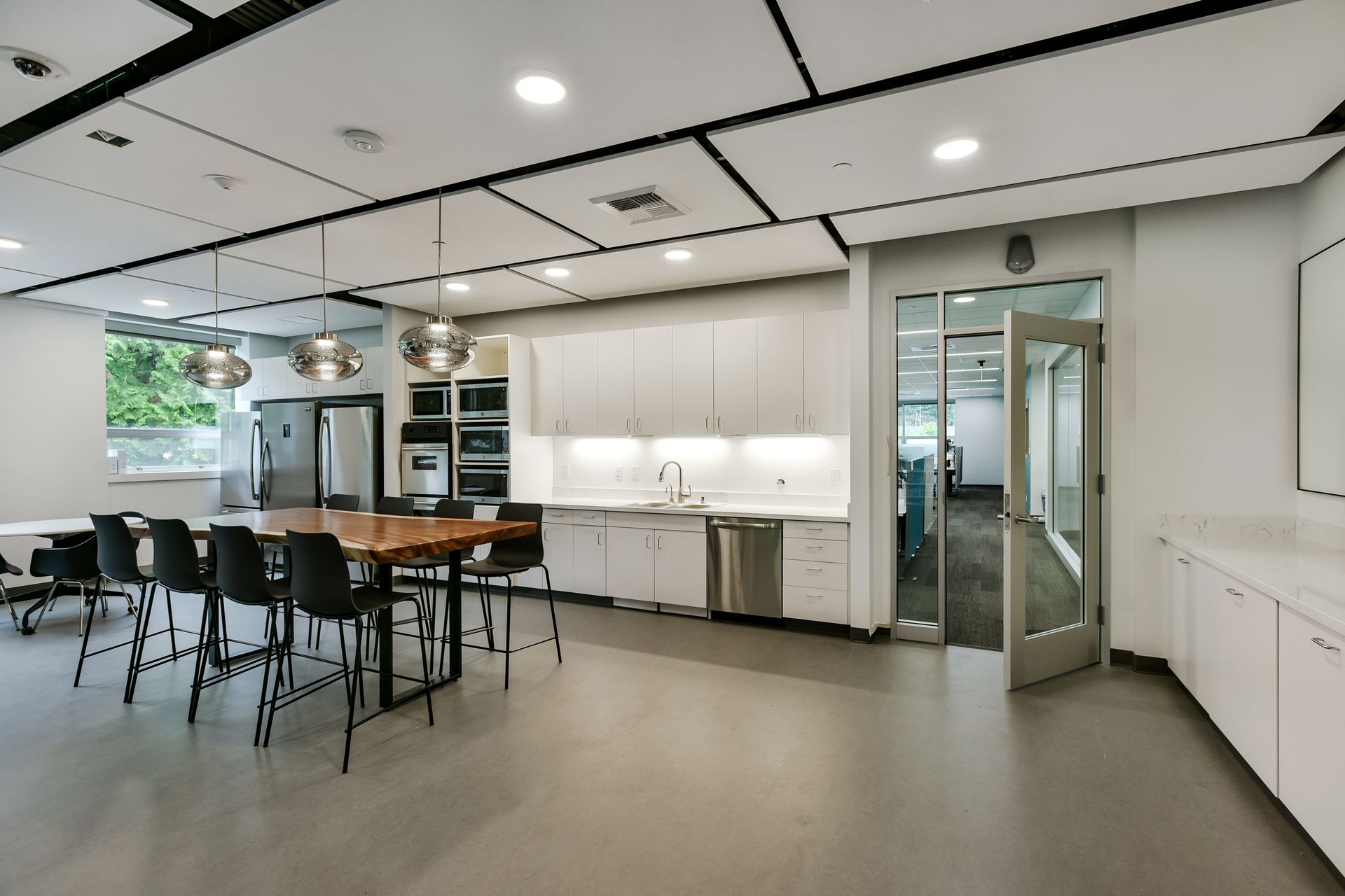 Modern office kitchen with long table, white cabinets, and stainless steel appliances.