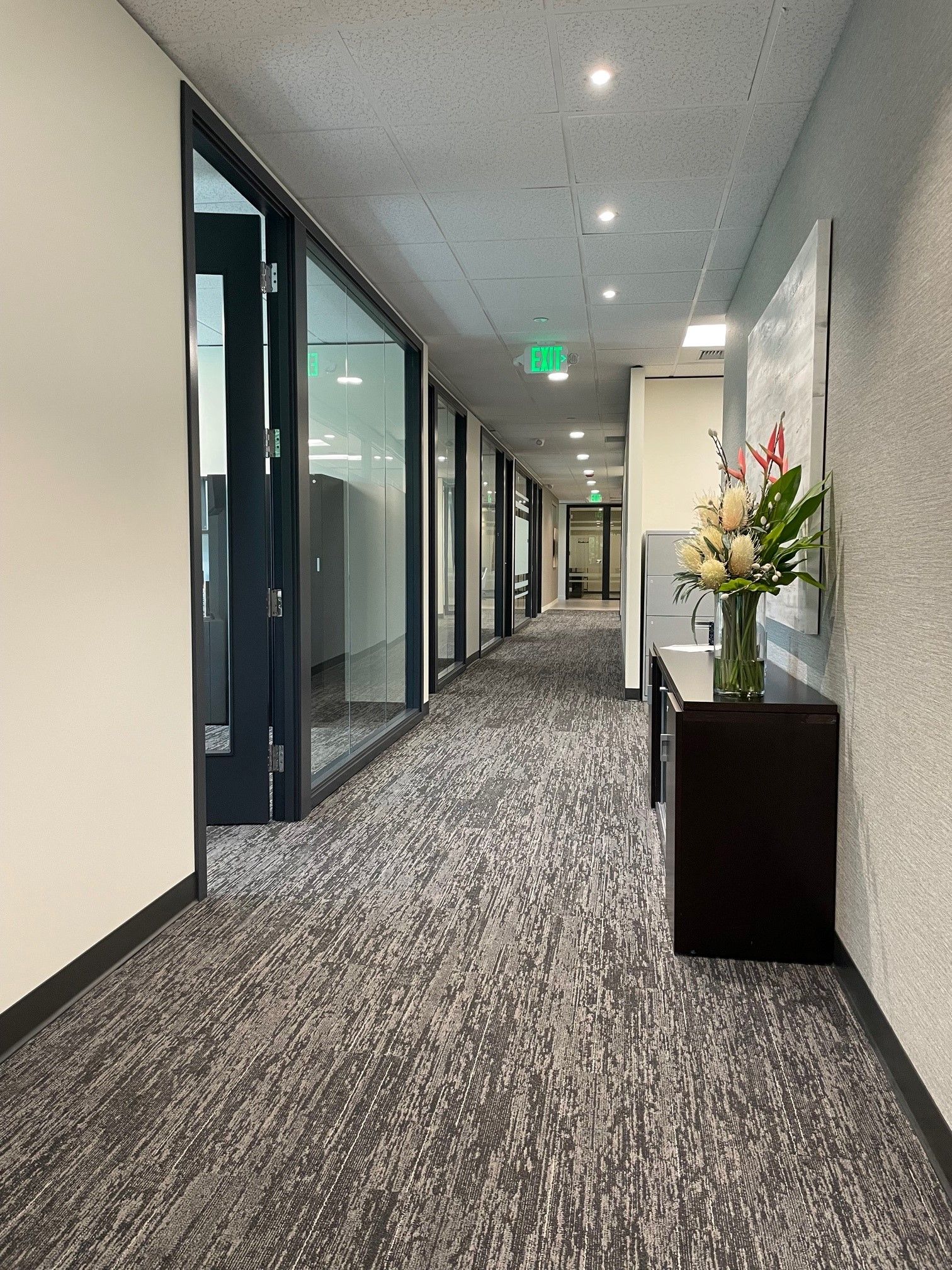 Hallway with glass-walled offices on the left, a dark console with flowers on the right, and patterned carpet.