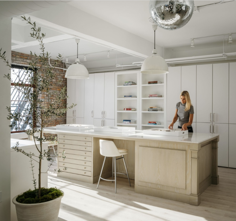 Woman at a large white worktable in a bright studio. White cabinets and a disco ball are also present.