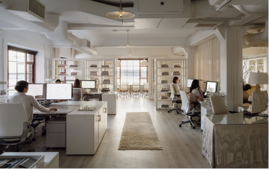 Bright, open office space with multiple desks, computers, and employees. White walls, wood floor, natural light.