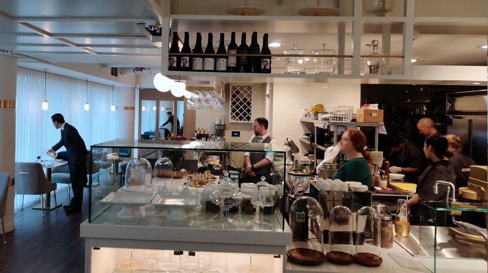 Modern cafe interior with people seated at tables, glassware on the counter, and bottles on shelves overhead