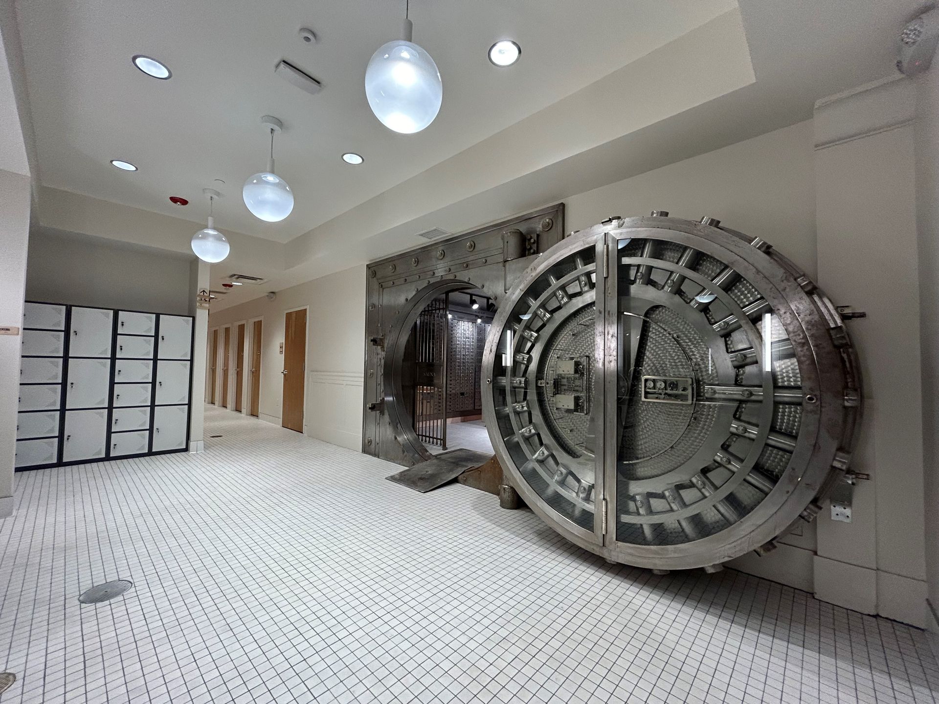 A large open bank vault door dominates a hallway. Lockers and doors line the left wall. White tiled floor.