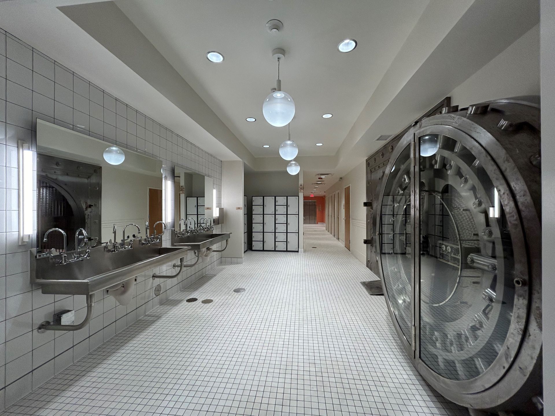 Hallway with vintage bank vault door, sinks, and tiled floor. White walls, round lights.