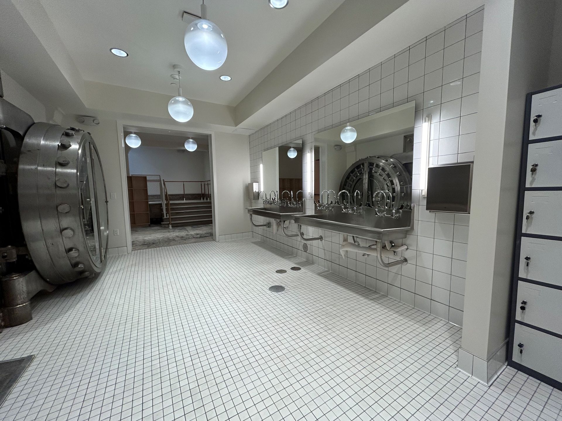 A bathroom with a large vault door. White tiled floor and walls. Sinks and mirrors. Lockers on the right.
