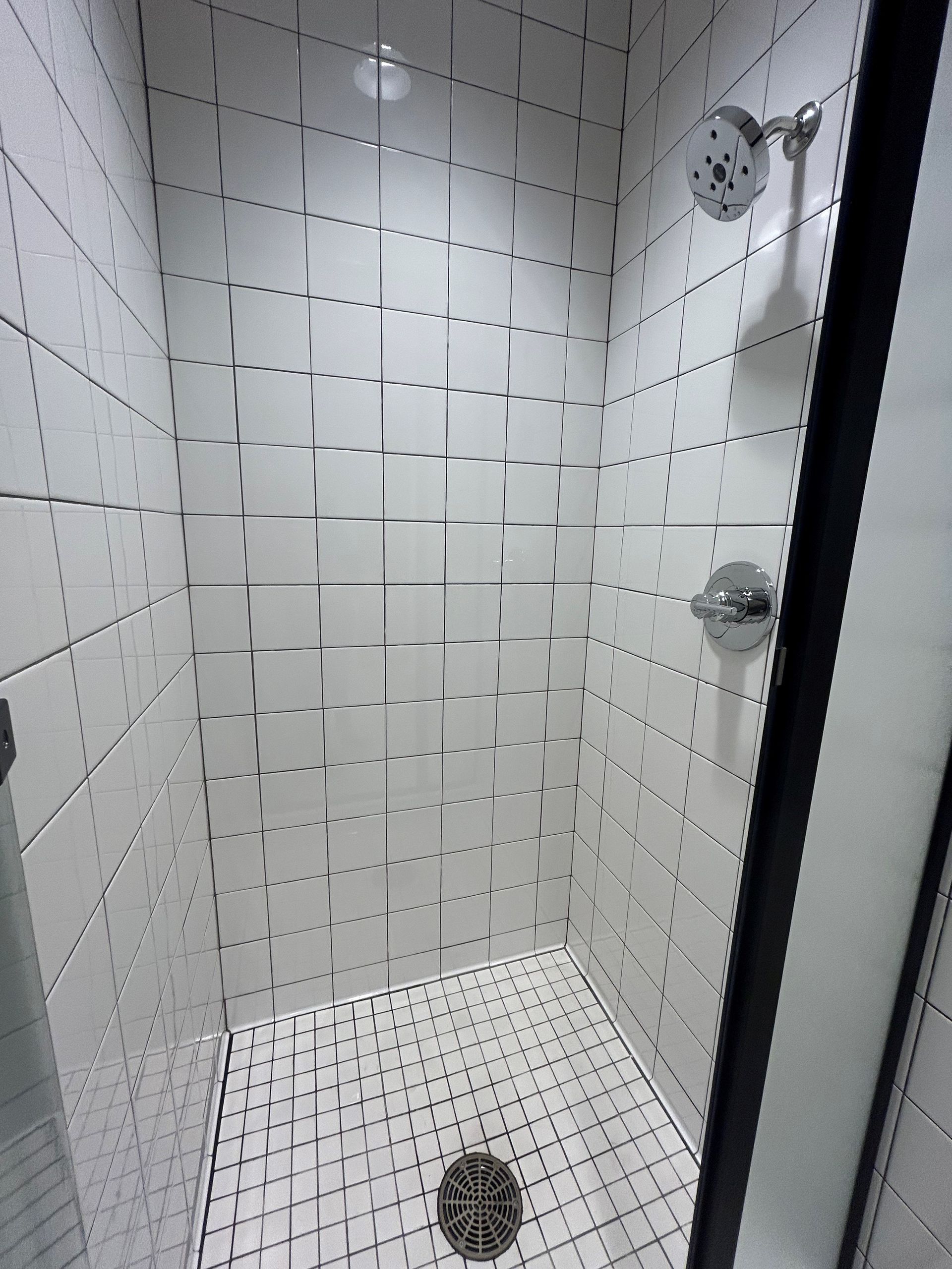 White tiled shower with a drain and fixtures; showerhead and water control knobs.