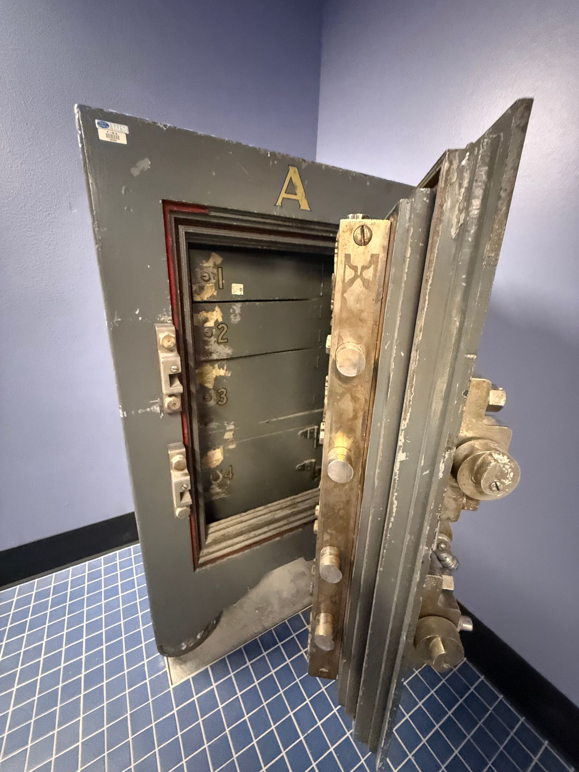 An open, old, gray bank safe door with exposed locking mechanisms, set against blue walls and a checkered floor.
