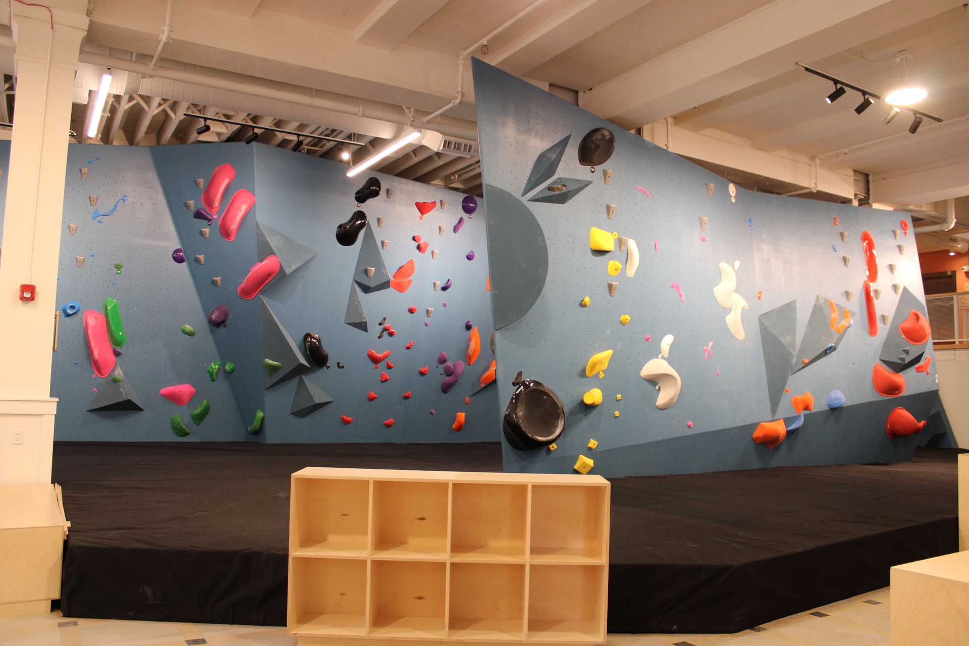 Bouldering wall with colorful climbing holds, indoors. A wooden storage unit sits in front.