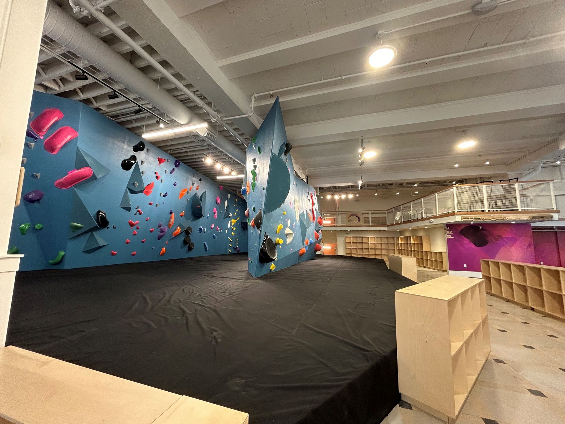 Indoor climbing gym with blue walls, colorful handholds, and black mats. Wooden storage and mezzanine.