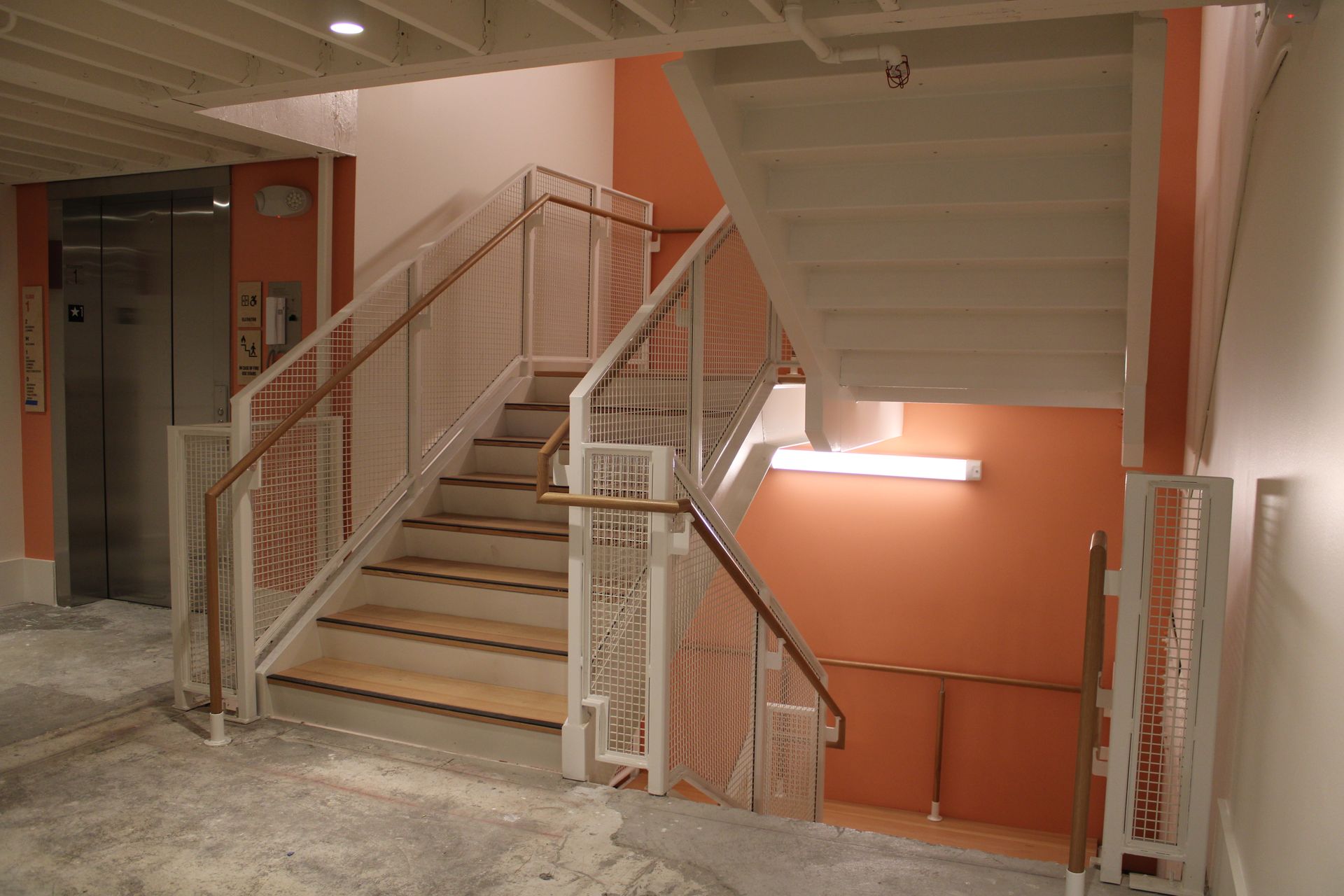 Interior view of stairs and an elevator with light orange wall. White railings and wooden steps.
