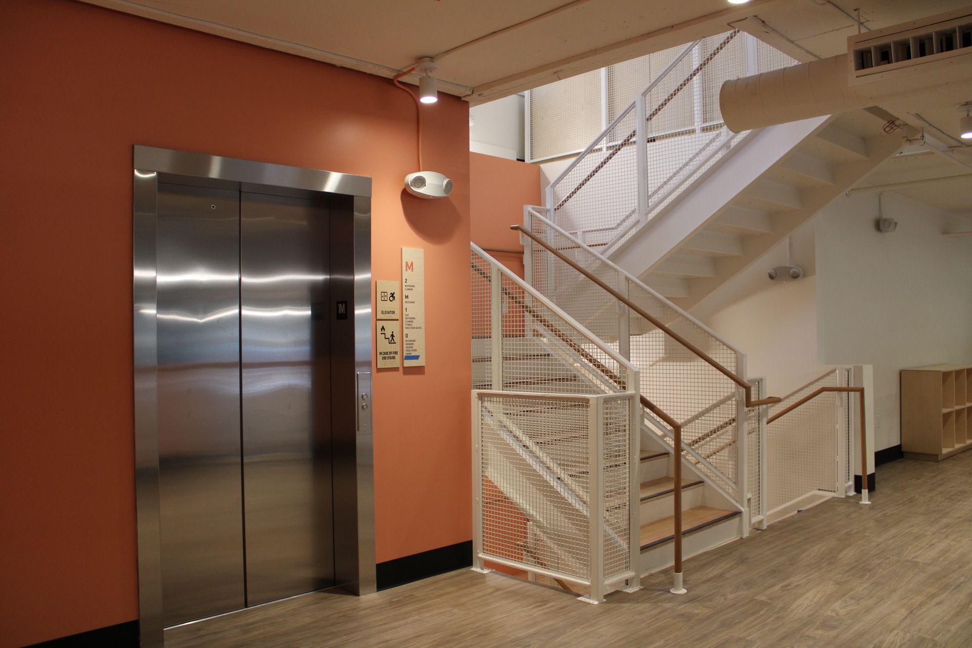 Elevator and staircase in a building, next to a salmon-colored wall. White railings and stairs.