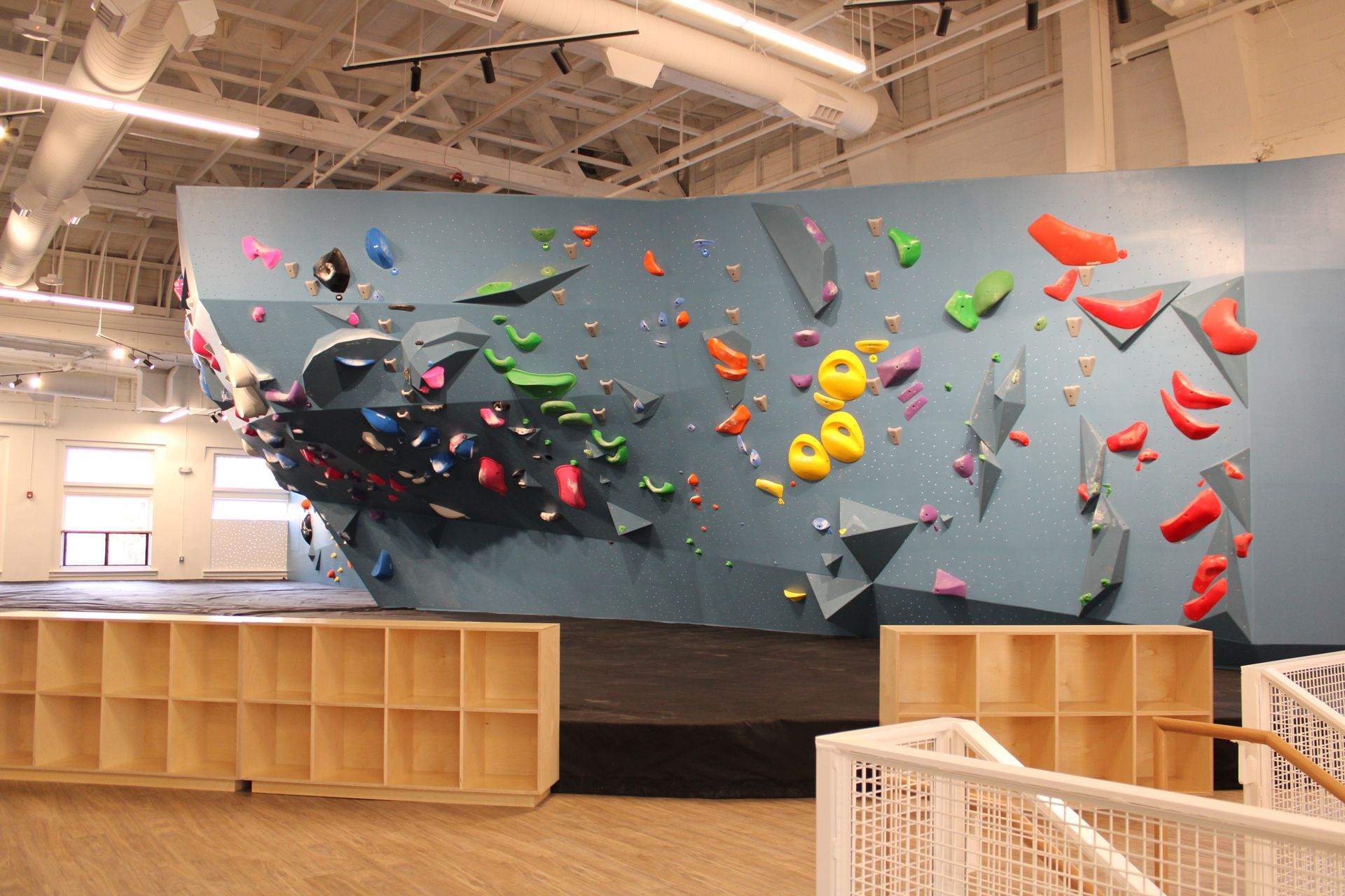 Indoor climbing wall with colorful holds. Wooden cubbies and stair rail in foreground.