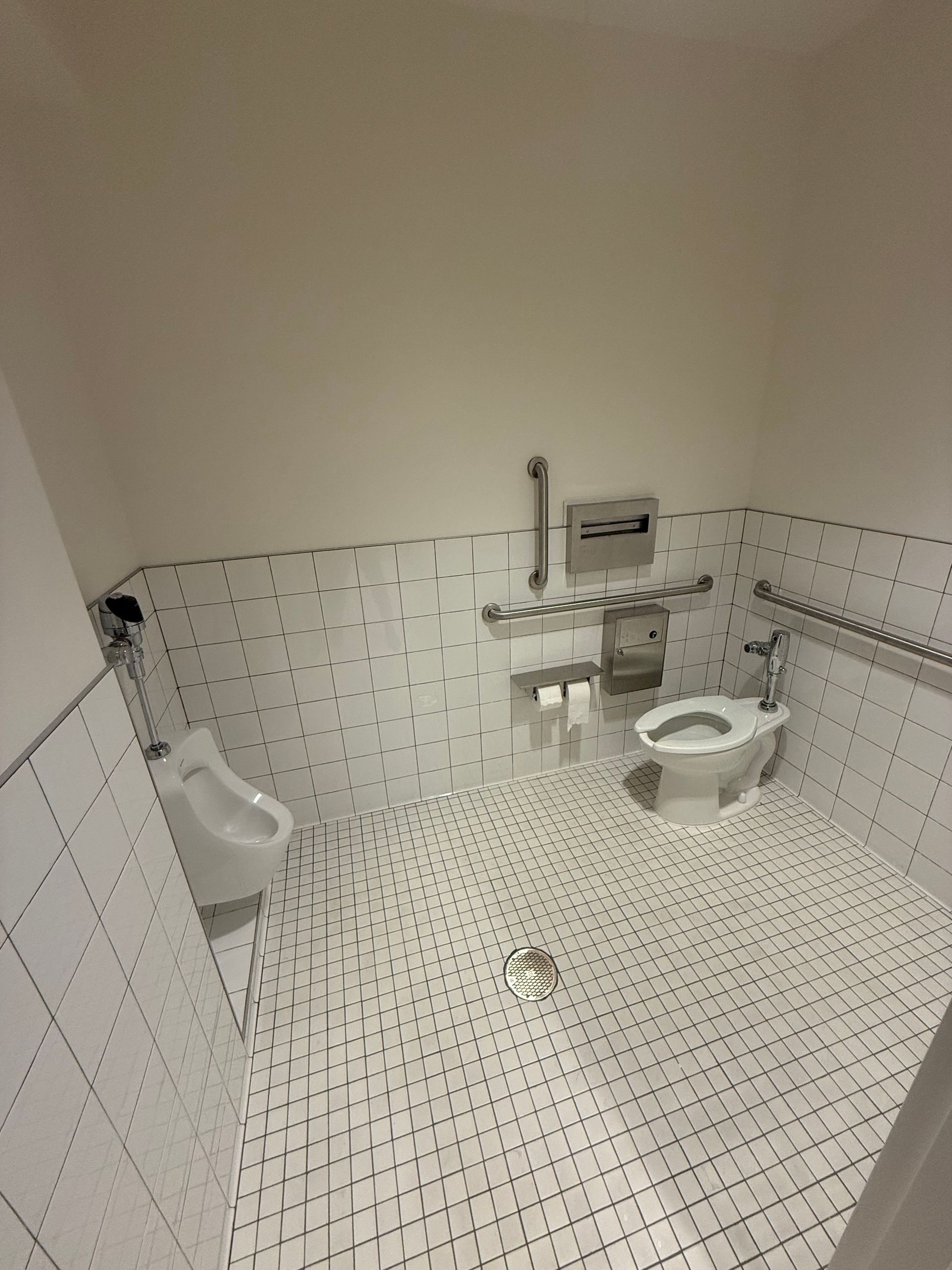 Public restroom with white tile walls and floor. Includes a urinal, toilet, and grab bars.