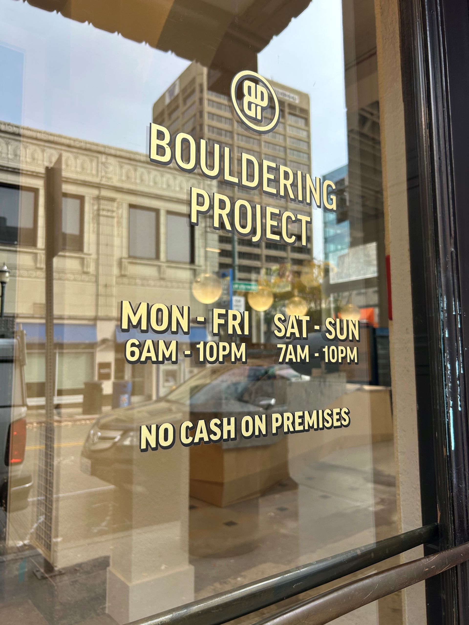 Sign on a glass door for Bouldering Project with hours and No Cash on Premises displayed.