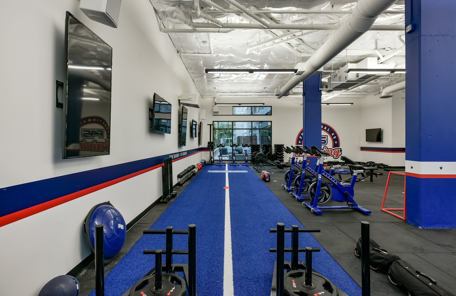 Gym interior with blue track, exercise equipment, and TVs on white and blue walls.
