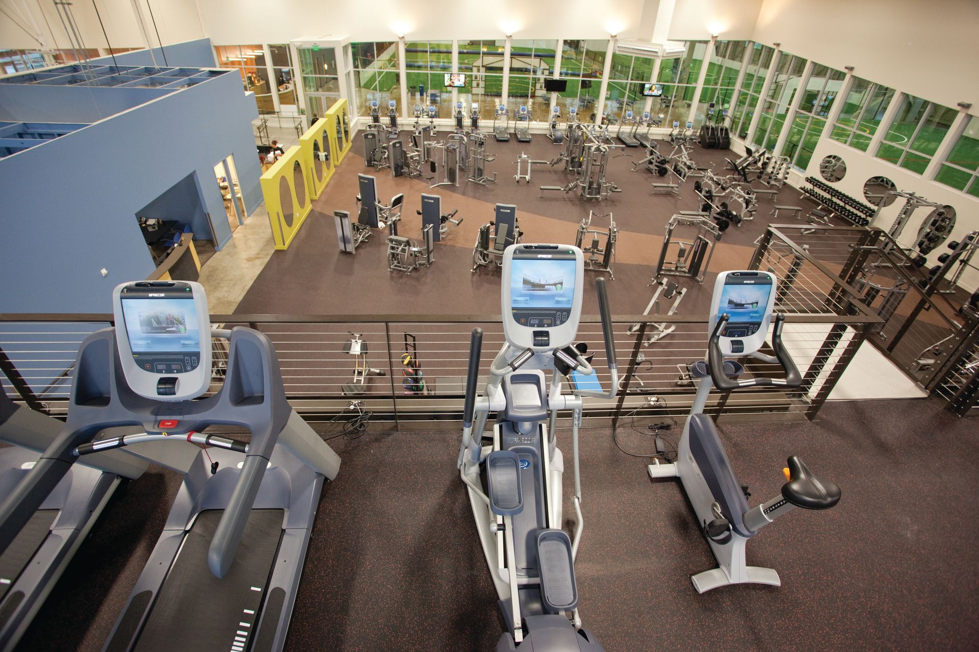 Gym interior with treadmills, elliptical, and exercise equipment. Large windows and black flooring.