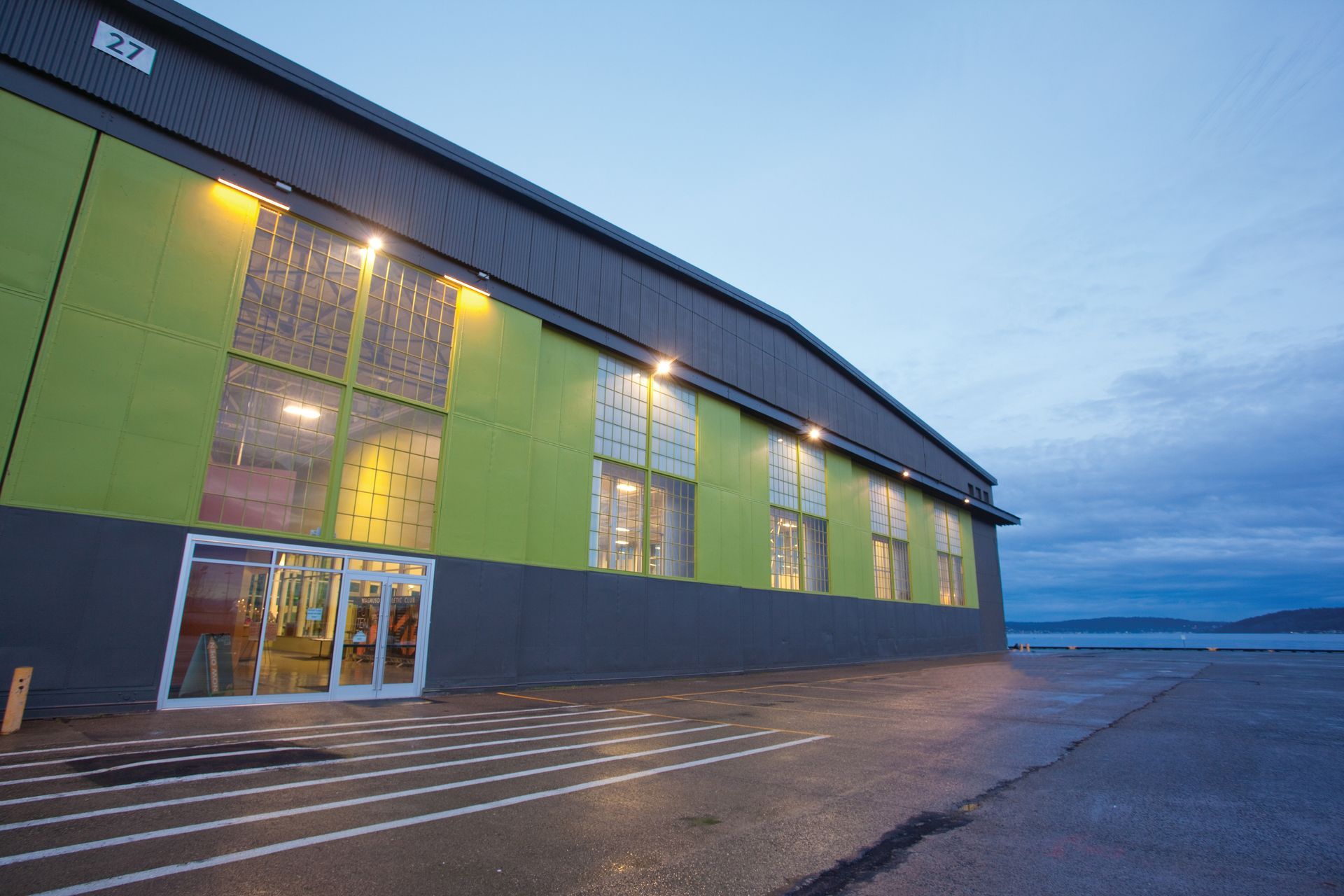 Large industrial building with green and grey panels, illuminated at dusk. Glass doors and windows visible.