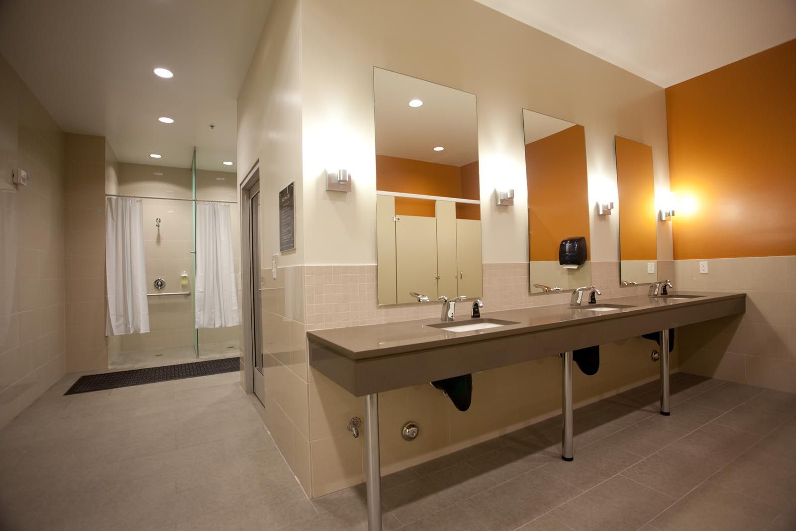 A public restroom with sinks, mirrors, stalls, and a shower area. Beige and orange walls, gray floor.